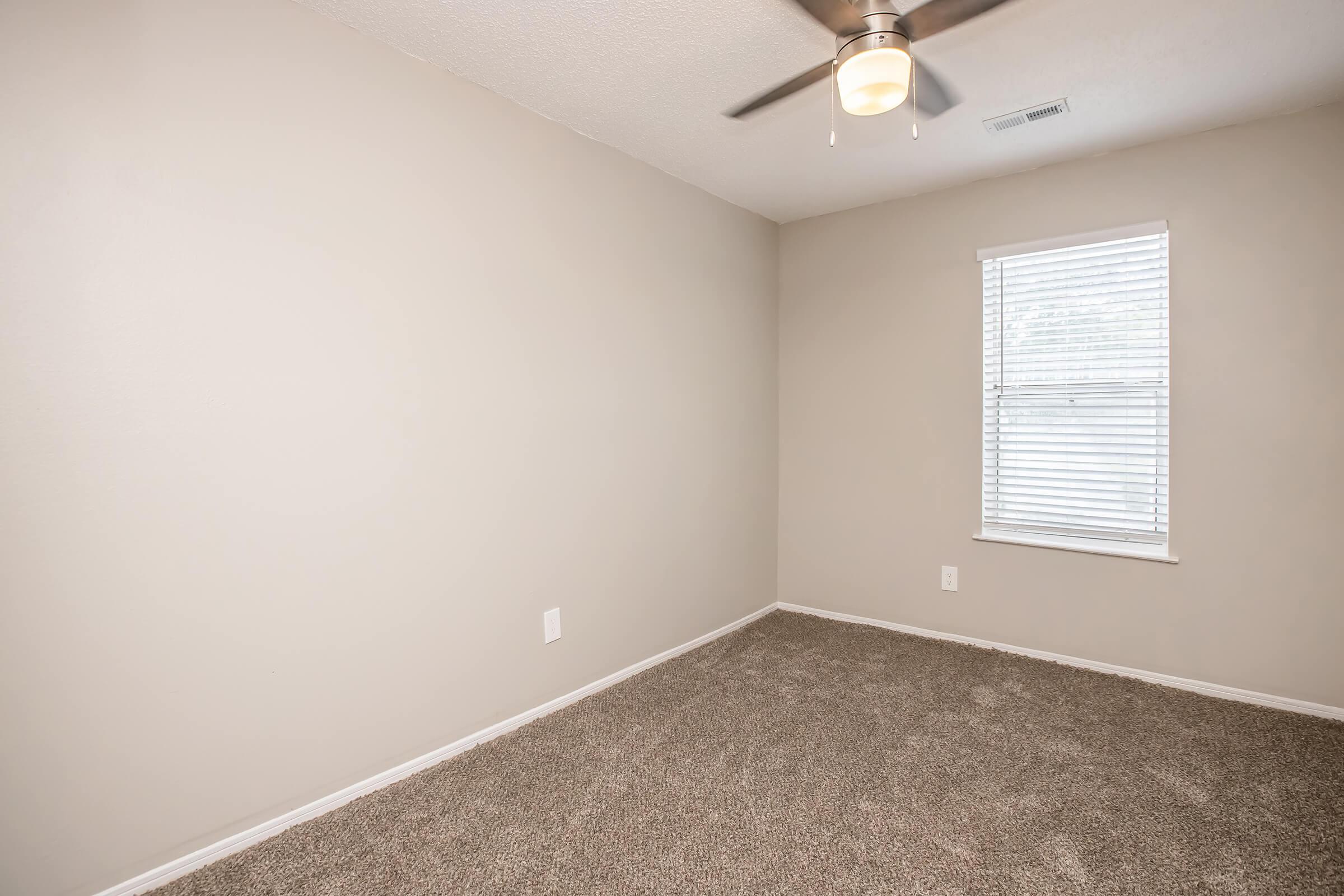 A small, empty room with beige walls and a carpeted floor. There is a ceiling fan above and a window with white blinds letting in natural light. The space is uncluttered, highlighting its potential for various uses or furnishings.
