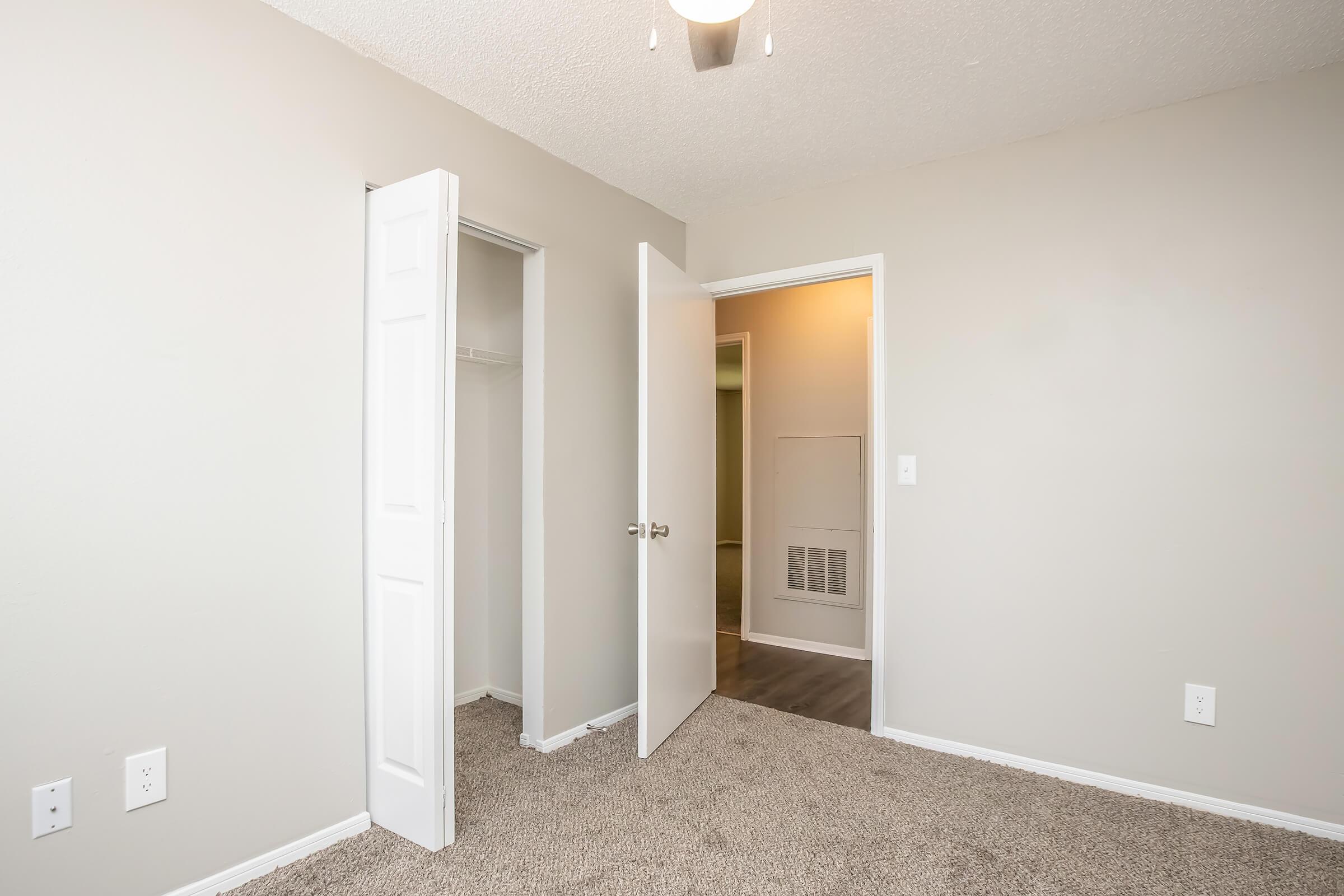 A clean and simple room with beige walls and carpet. Two white doors are open, one leading to a closet and the other to another space. A light fixture on the ceiling provides illumination. The room appears well-maintained and inviting, suitable for various uses.
