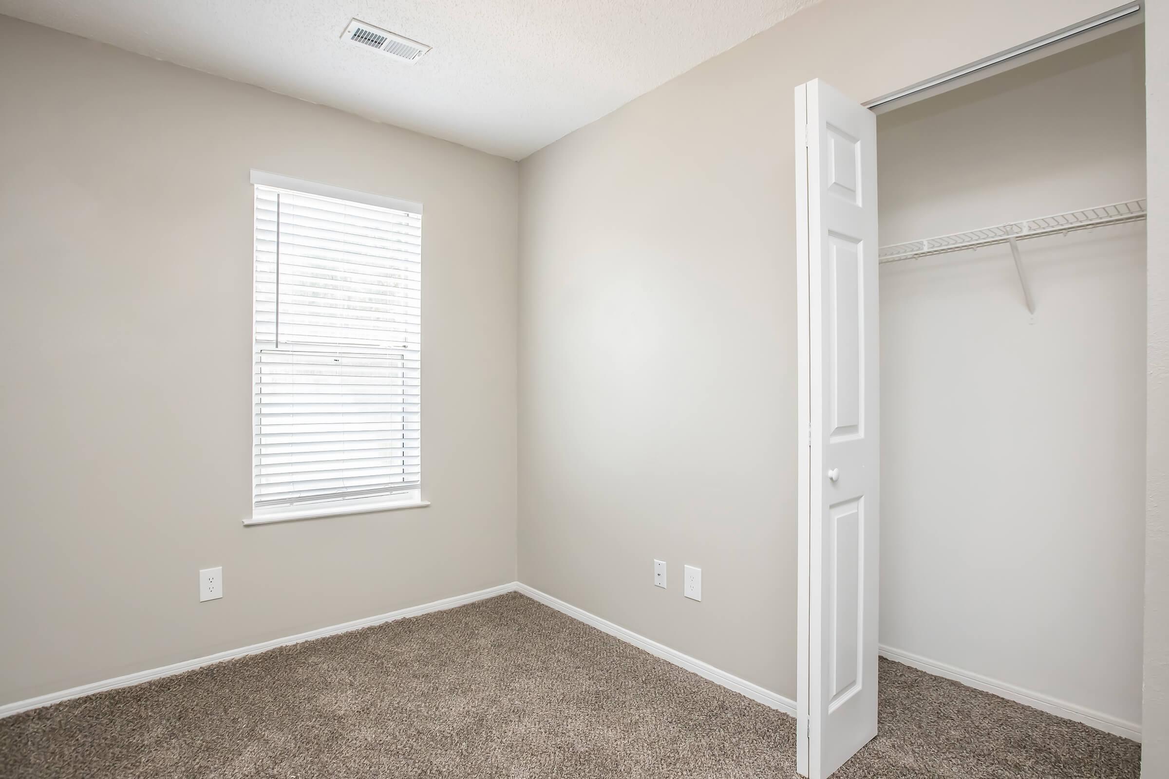 A vacant room with light beige walls and carpet. The room features a window with white blinds, allowing natural light to enter. An open closet with a white door is visible on the right side of the image. The overall space appears clean and minimalistic, suitable for various uses.