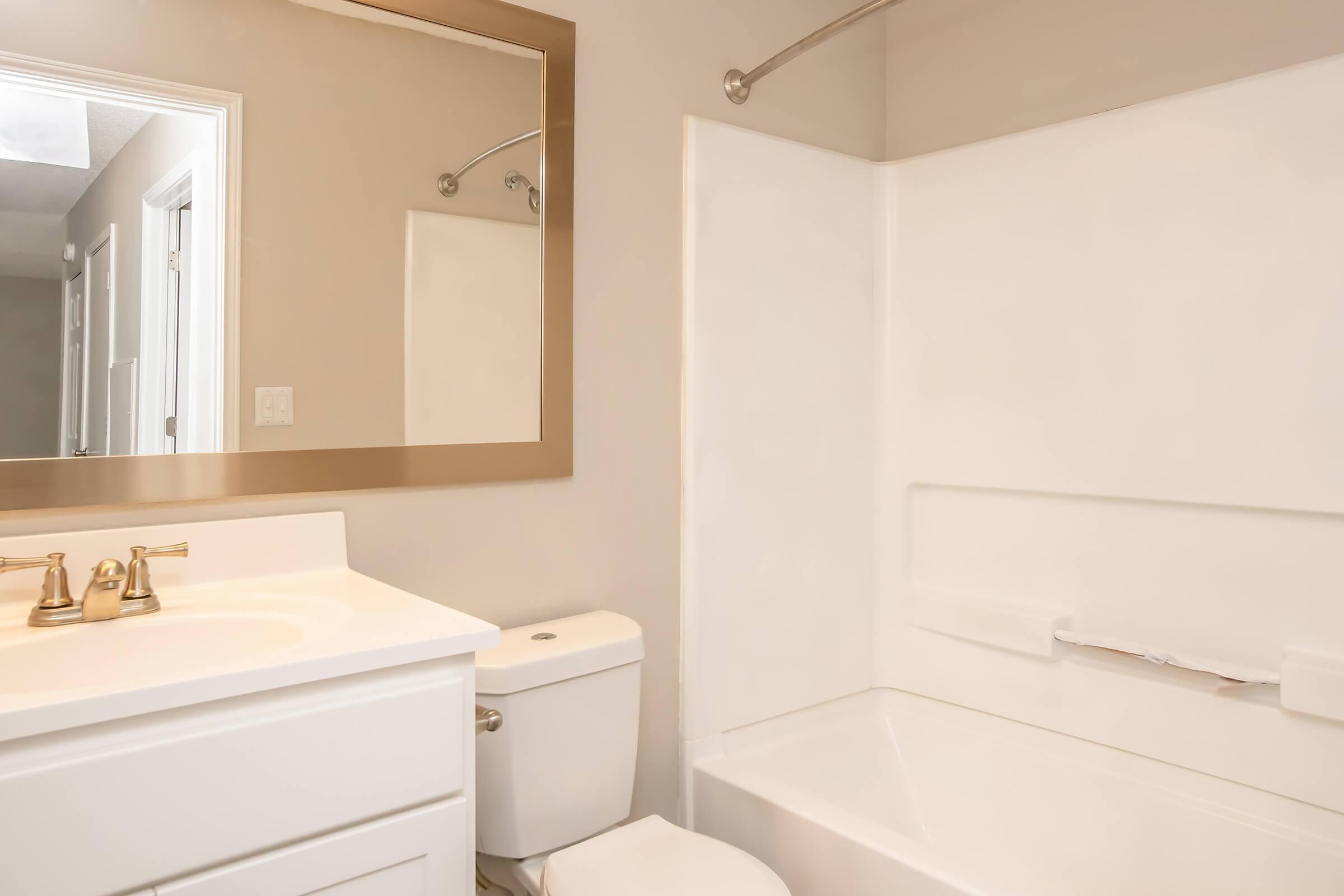 A modern bathroom featuring a white bathtub and shower, a white vanity with a sink and gold-colored faucets, a toilet, and a large mirror above the sink. The walls are painted a light color, and there is a doorway visible in the background.