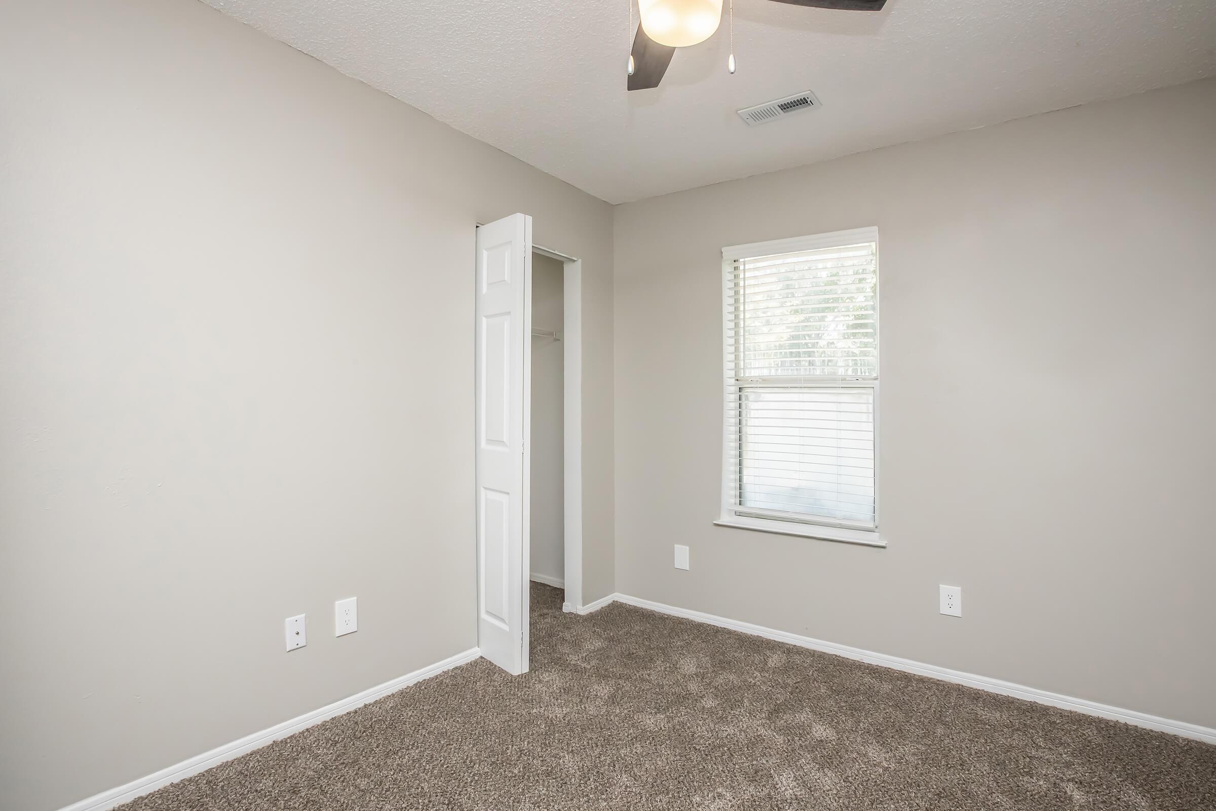 A vacant, well-lit bedroom featuring light gray walls, carpeted flooring, a ceiling fan, and a window with white blinds. There's a closet with a partially open door on one side, providing a sense of space and simplicity in the room's design.