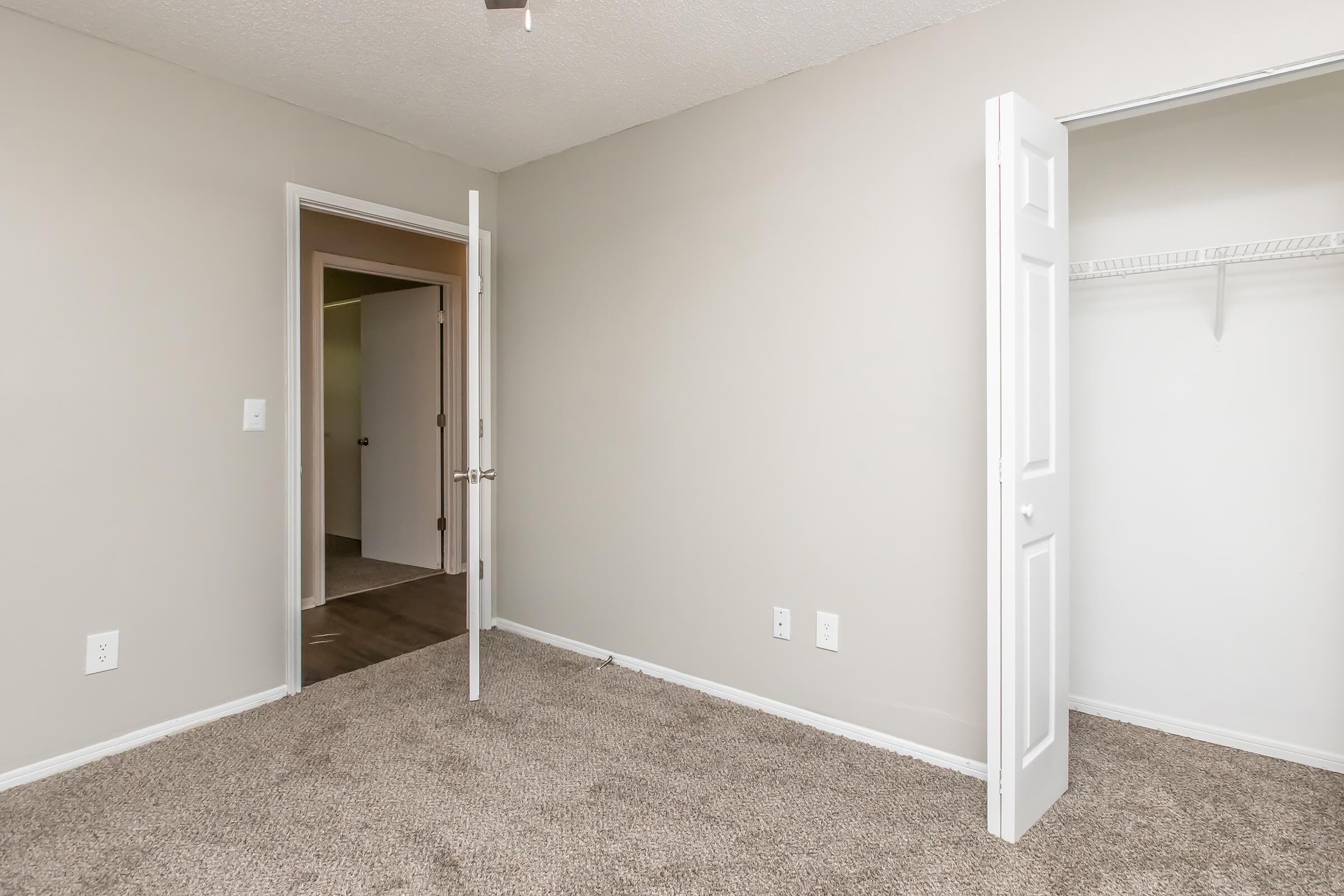 Empty room with light gray walls and beige carpet. A half-open white door leads to another room, while a closet with a hanging rod is visible on the right. The room is well-lit, creating a spacious feel.