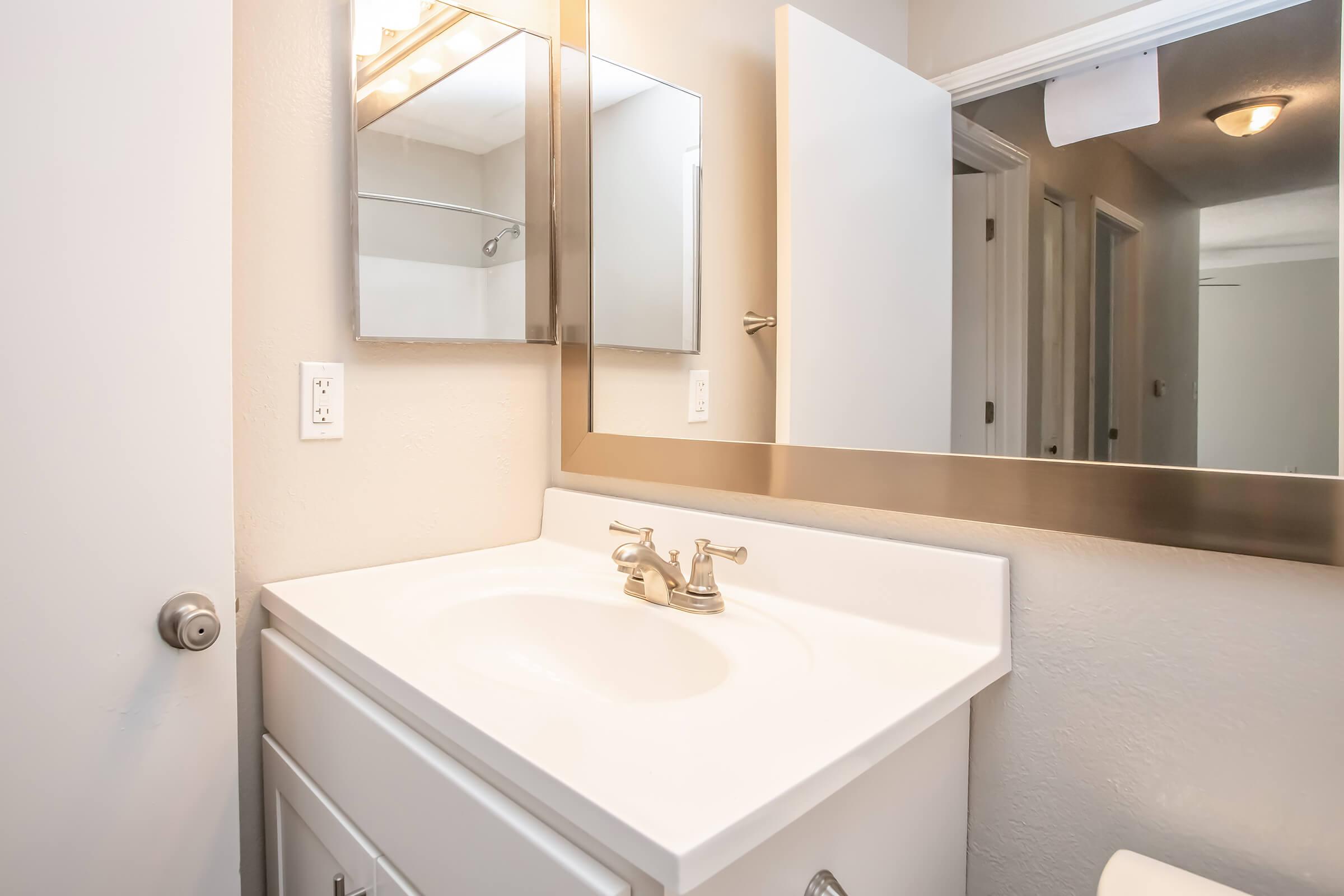 A clean bathroom with a white vanity featuring a sink and chrome faucets. A large mirror above the sink reflects the light-colored walls. There are doors to the right leading to other rooms, and a shower area is partially visible in the background. The overall ambiance is bright and uncluttered.