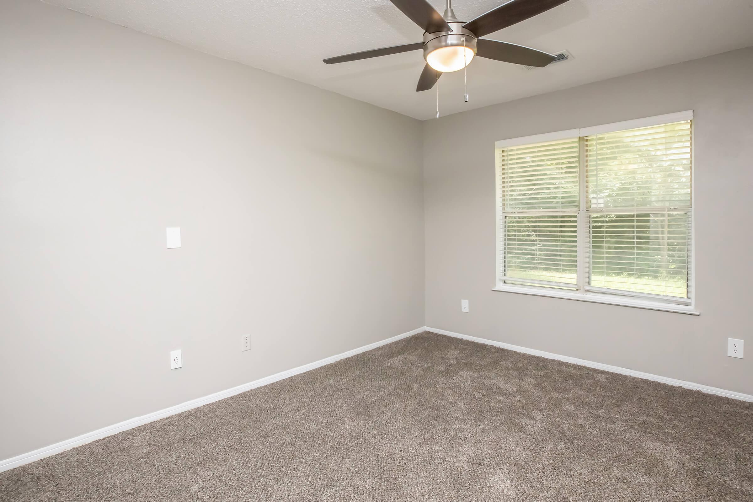 Empty room with light gray walls and a ceiling fan. The floor is covered in beige carpet. A window with blinds allows natural light to enter, showing greenery outside. The space appears clean and minimalistic, ideal for various uses such as a bedroom or office.