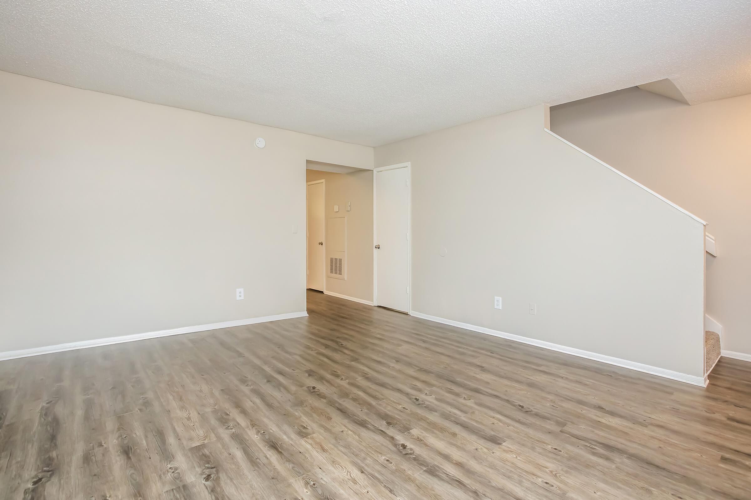 A spacious, empty living room with light-colored walls and a large window. The flooring is a modern wood laminate. A staircase leads to a second level in the corner. There is a doorway visible, suggesting access to other areas of the home. The room is well-lit and has a clean, neutral aesthetic.