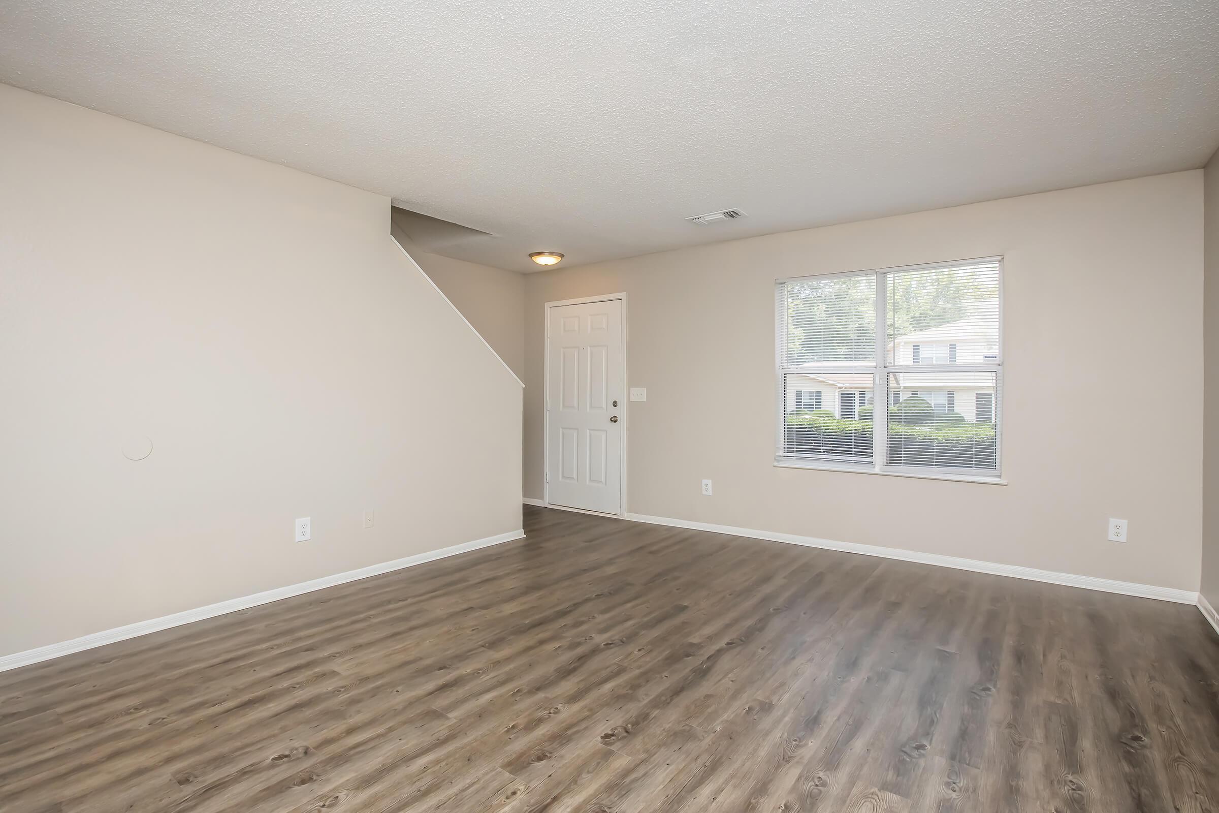 Spacious interior of a living room featuring hardwood-style flooring, a white door, and a large window with natural light. A staircase is visible in the background, and the walls are painted in a neutral color, creating a bright and open atmosphere.