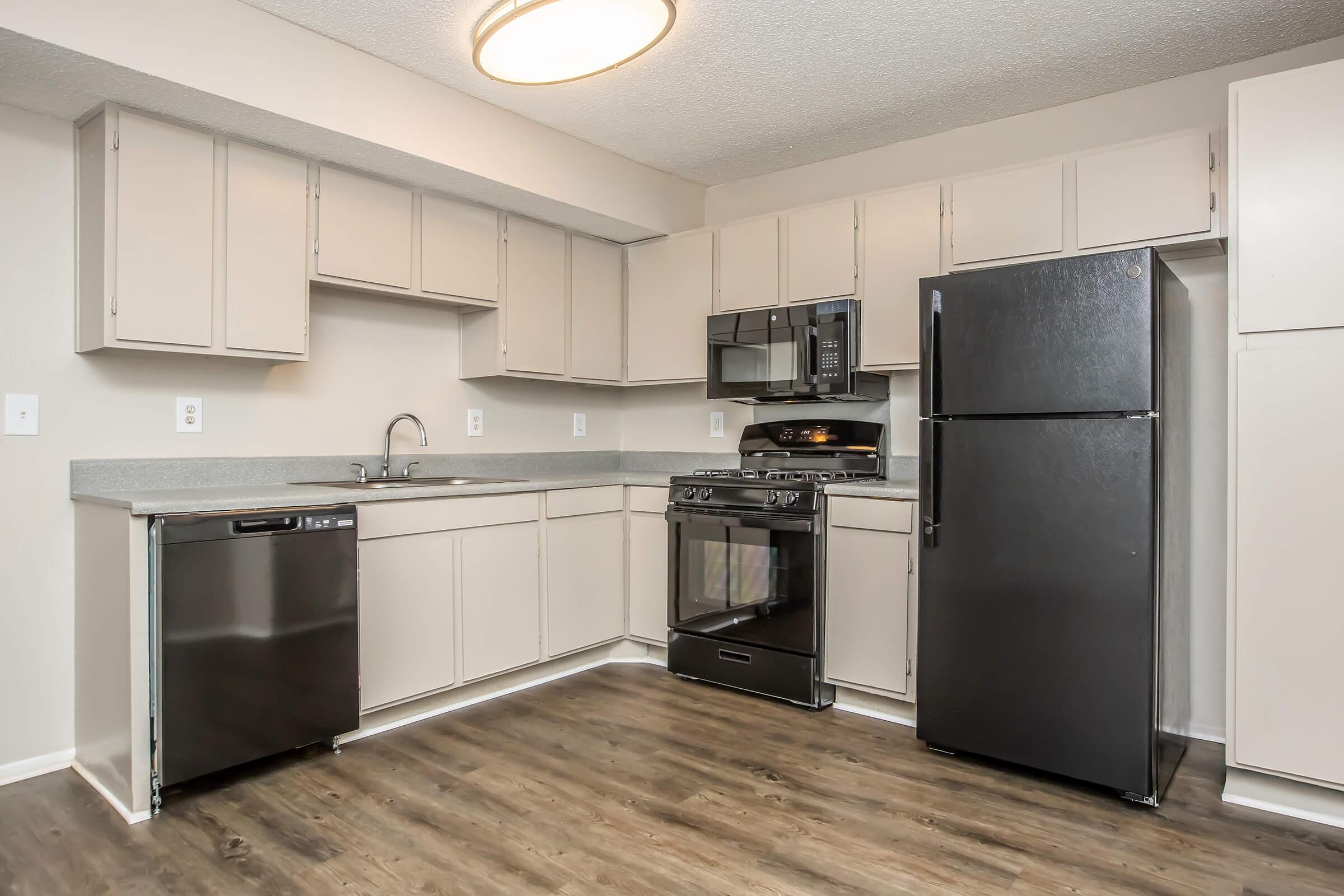 Modern kitchen featuring light-colored cabinets, a gray countertop, and stainless steel appliances. The kitchen includes a microwave, gas stove, and refrigerator, complemented by a black dishwasher. The flooring is a light wood laminate, providing a clean and contemporary look.