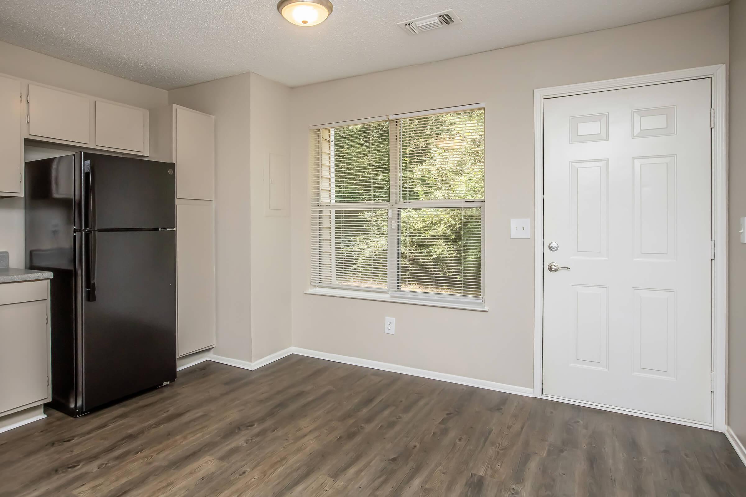A small kitchen area featuring a black refrigerator, light-colored cabinets, a window with blinds allowing natural light, and a door leading outside. The flooring is dark, and the walls are painted a neutral color, creating a cozy and inviting atmosphere.