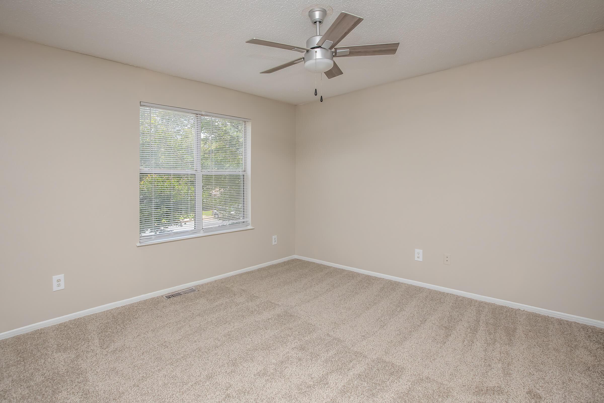 Empty room with beige walls and light-colored carpet. A ceiling fan is mounted to the ceiling, and there is a window with blinds that allows natural light to enter. No furniture is present in the space, creating a clean and simple environment.