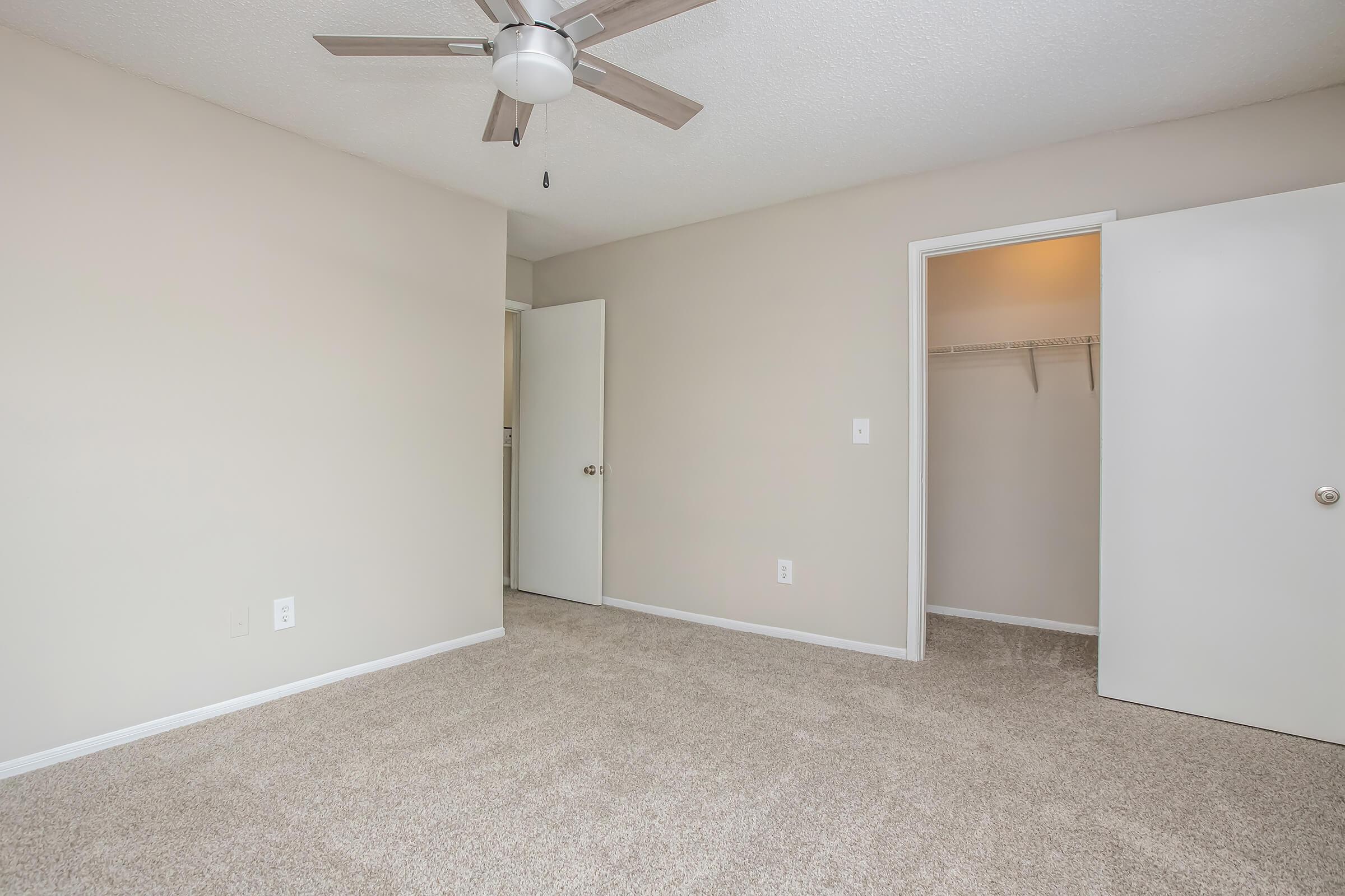A spacious, unfurnished room with light beige walls and newly installed carpet. It features a ceiling fan and an open closet space with a sliding door. The room has one door leading to a hallway and another door leading into a closet. The overall ambiance is calm and inviting, perfect for personalizing.