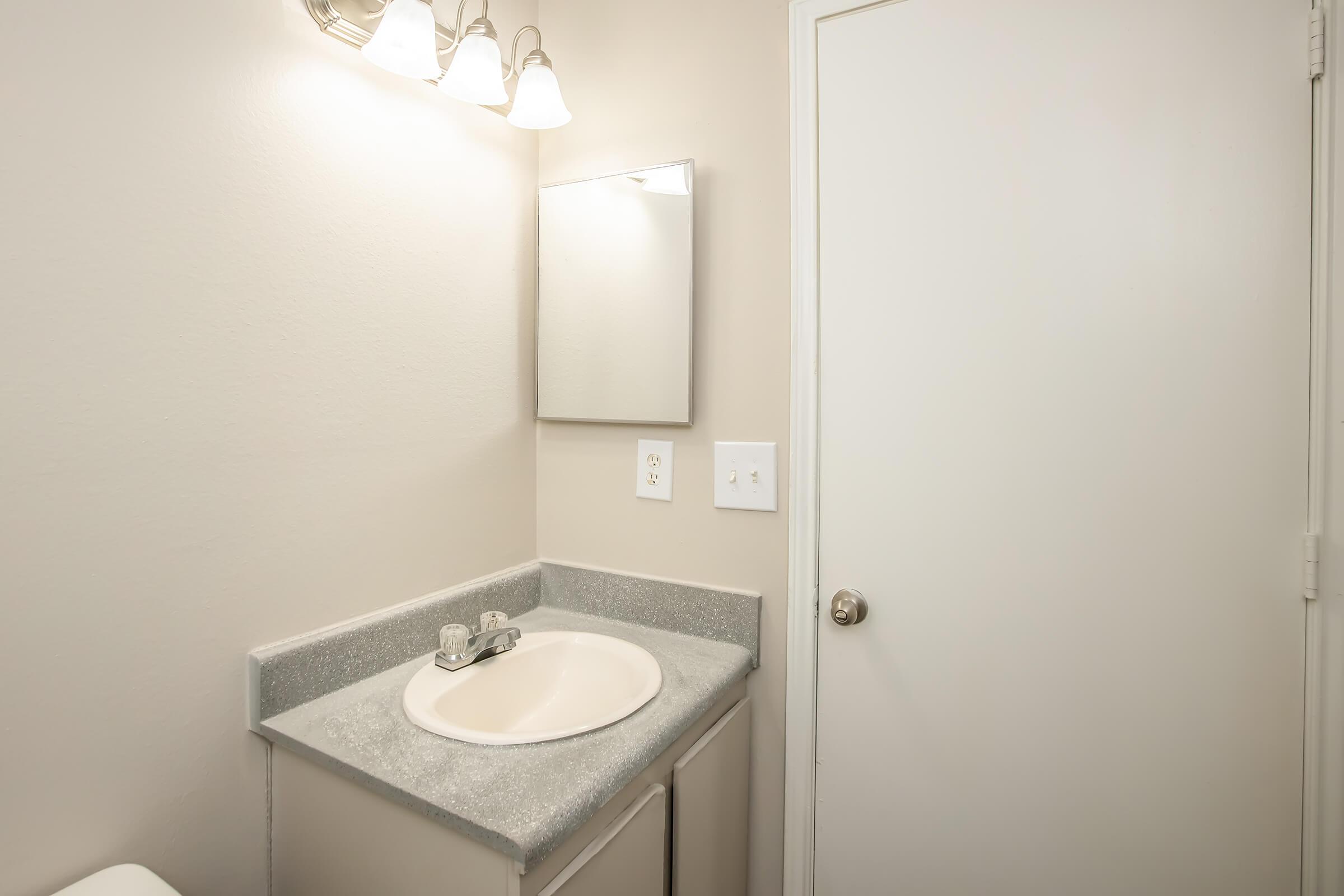 A simple bathroom scene featuring a gray countertop with a sink, a wall-mounted mirror above the sink, and light fixtures above the mirror. The walls are painted a light color, and there is a closed white door visible on the right side of the image.