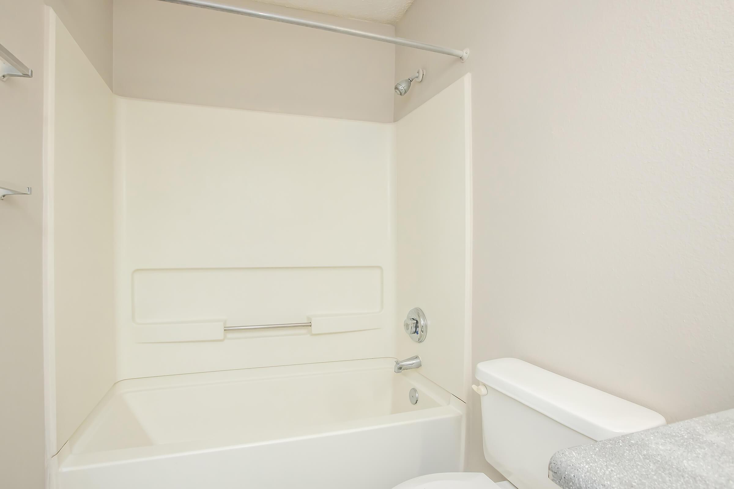A clean, minimalist bathroom featuring a white bathtub with a grab bar, a white toilet, and smooth beige walls. The space has a simple design, with no decorative elements, giving it a modern and fresh appearance.