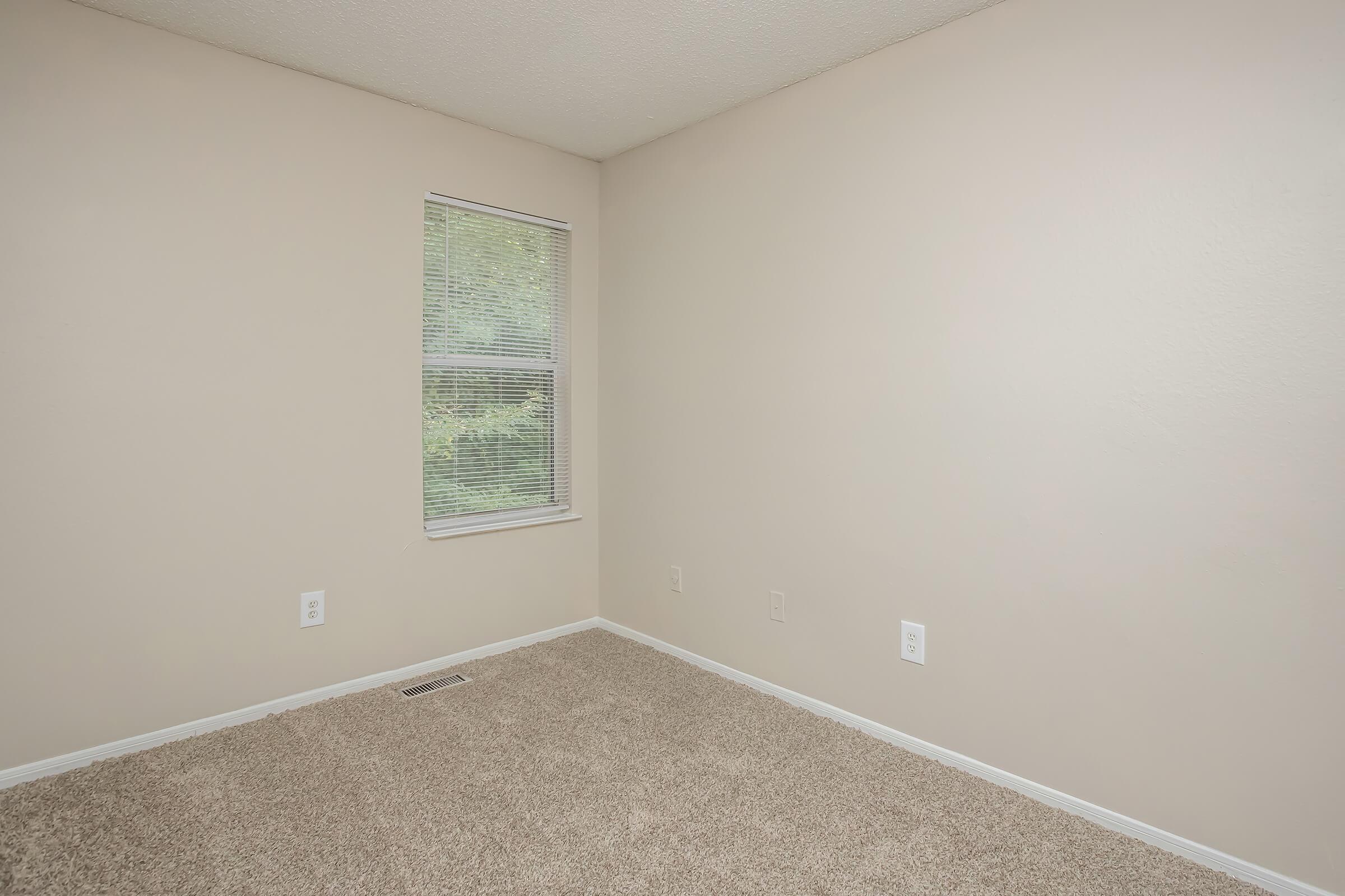 A small, empty room with light beige walls and beige carpet. There is a single window with white blinds on one wall, allowing natural light to enter. The room has no furniture or decorations, creating a minimalist and spacious appearance.