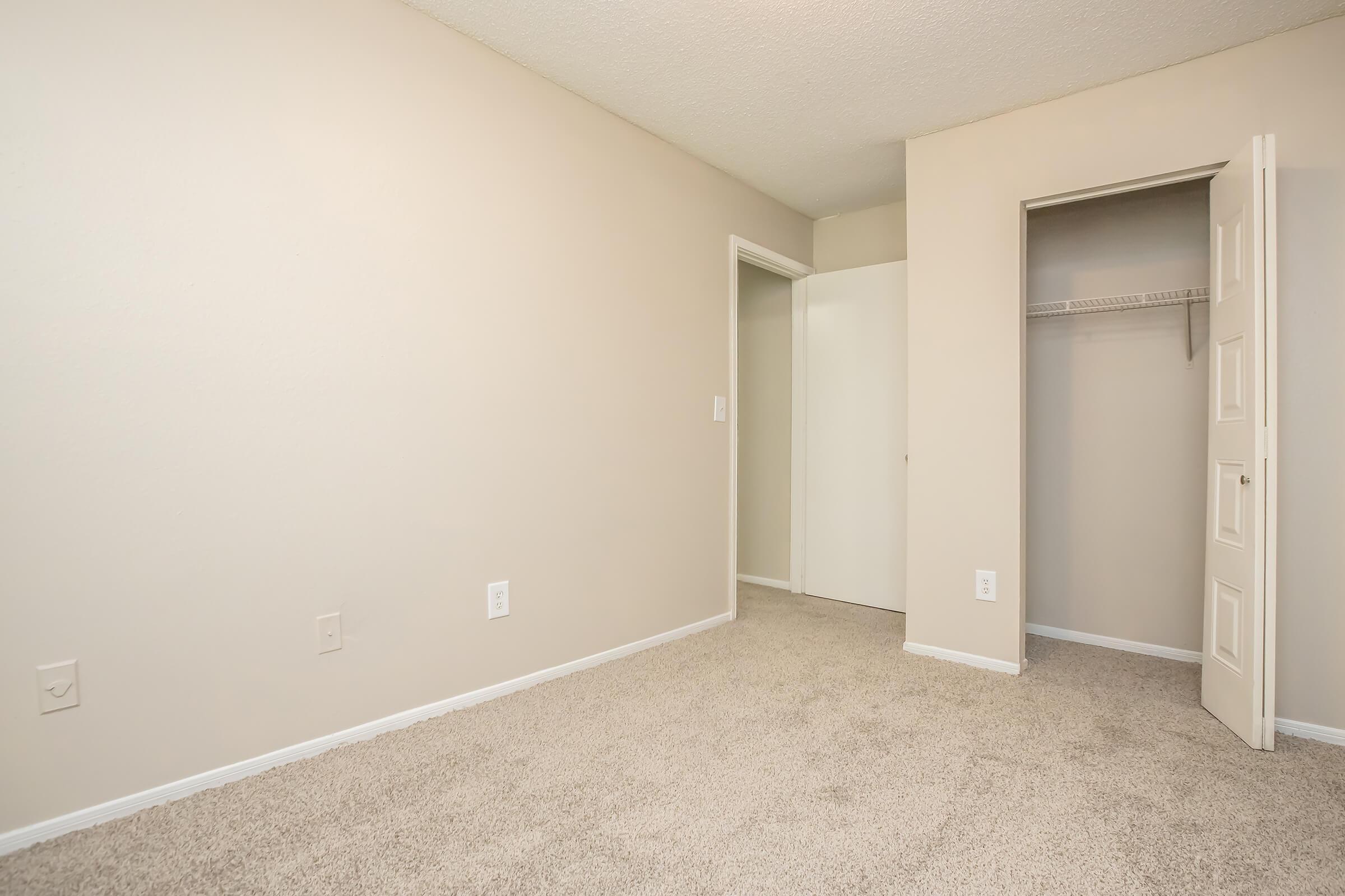Empty room with beige walls and carpeted floor. One wall features a closet with an open door, and there are two additional doorways leading to other areas. The space is well-lit and has a clean, minimalistic appearance, suitable for various uses such as a bedroom or office.