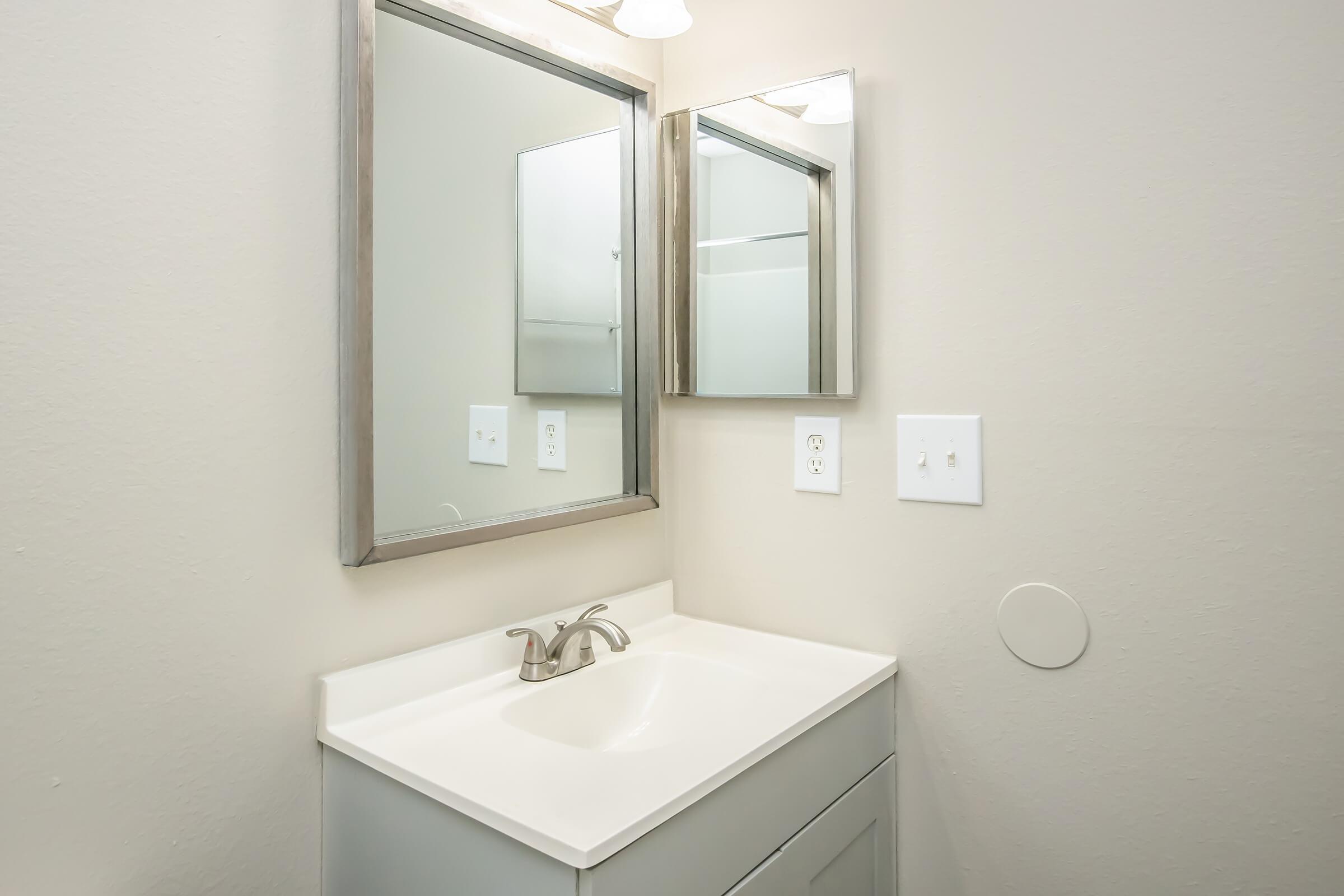 A clean bathroom sink with a white countertop and a rectangular mirror above. The walls are a soft neutral color, and there are light switches and a power outlet visible nearby. The overall design is modern and minimalistic.