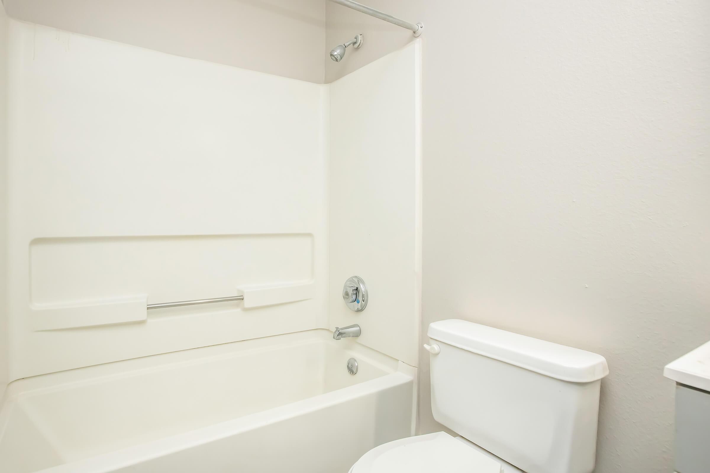 A clean and simple bathroom featuring a white bathtub with a showerhead, a toilet, and neutral-colored walls. The space is well-lit and has a modern appeal, with a focus on functionality and minimalism.
