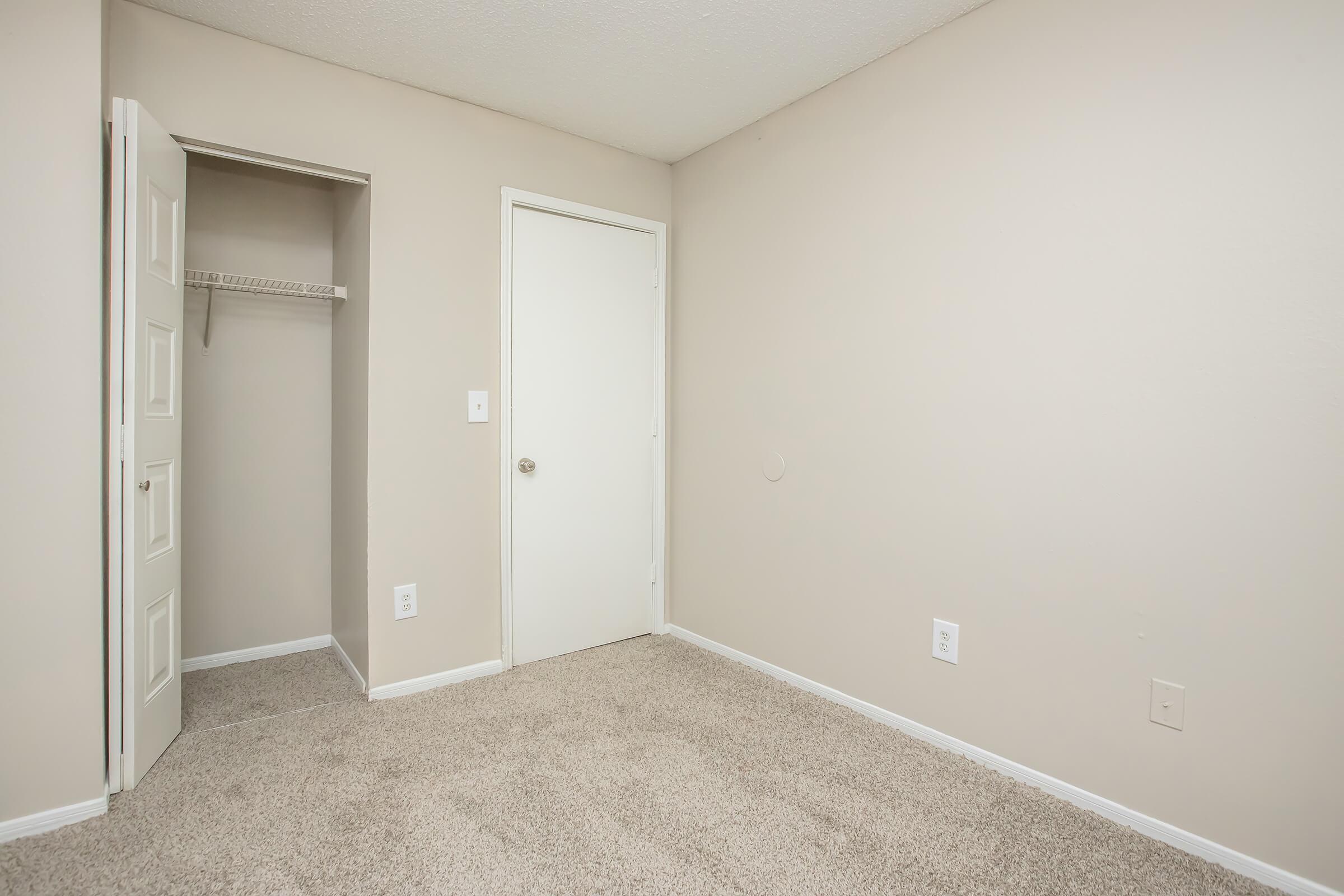 Empty room with light beige walls and carpeted floor. There's a closet on the left with a door ajar and another door leading to an adjacent space. The overall atmosphere is neutral and unfurnished, suggesting it could be a bedroom or office space.