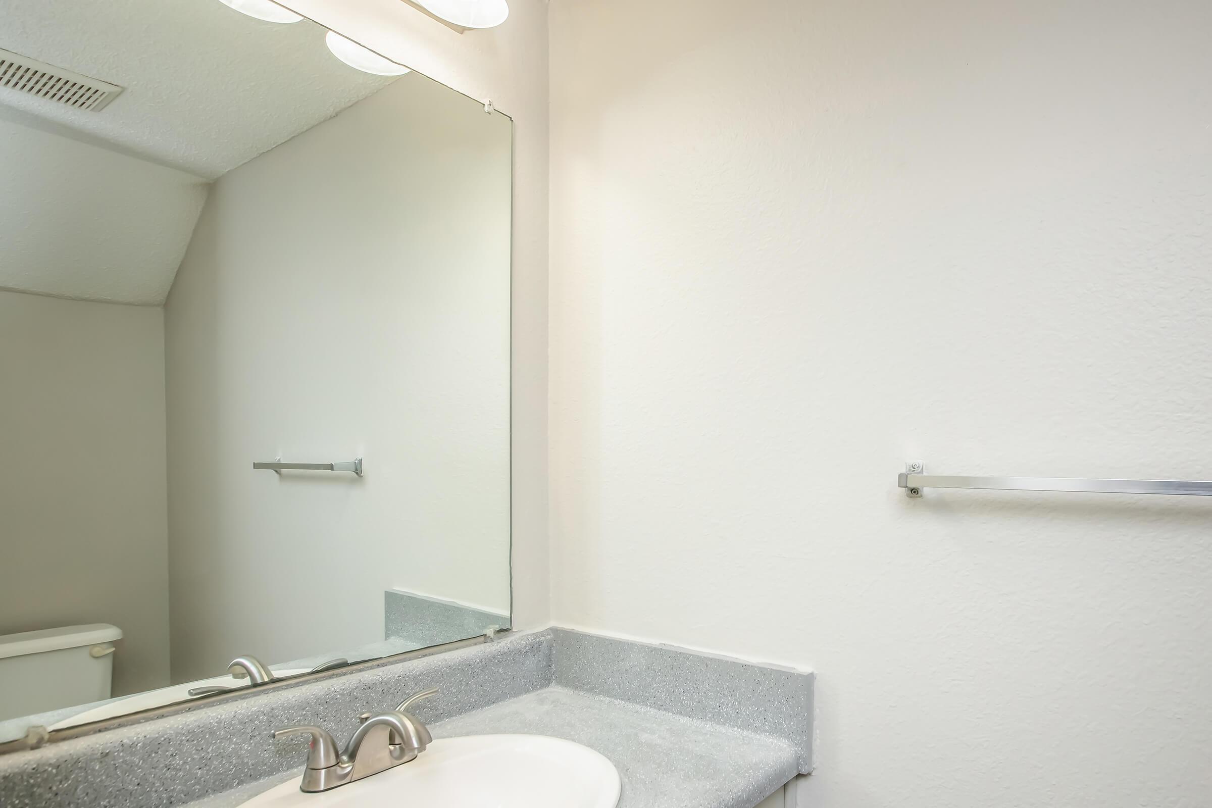 A clean, well-lit bathroom featuring a simple sink with a chrome faucet, a gray countertop, and a large mirror above. The wall is painted a light color, and there's a towel rack and a toilet visible in the reflection. The overall atmosphere is bright and minimalistic.