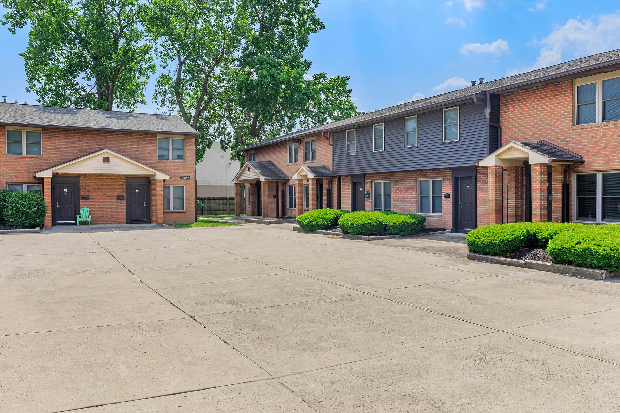 A view of a residential building complex featuring two brick buildings, one light-colored and one dark-colored, with a shared parking area. Lush green bushes line the pathways, and a single green chair is visible beside one of the entrances. The sky is clear with a few clouds.