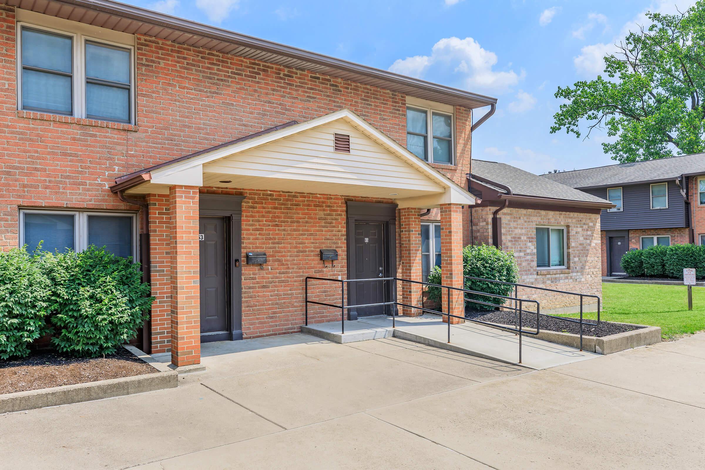 A brick apartment building with a light-colored entrance and dual doors under a peaked roof. It features a wheelchair-accessible ramp leading up to the entryway. Lush green shrubs line the front, with a grassy area on the side and another building visible in the background against a blue sky.