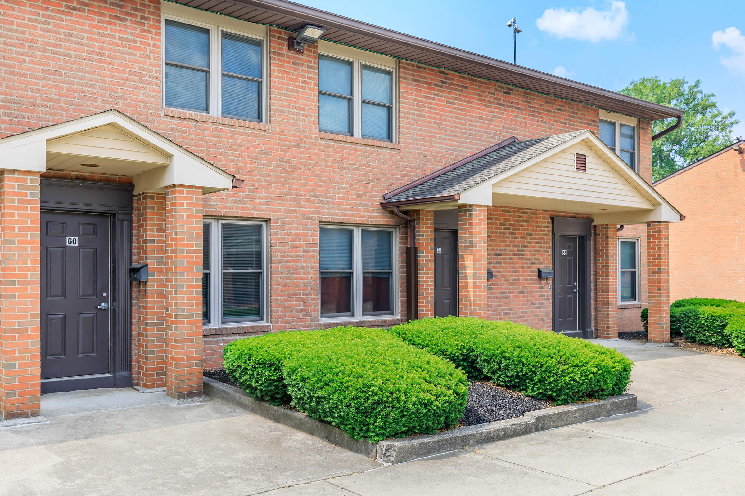 Two-story brick apartment building with multiple units. Each unit has a dark door and several windows. Lush green bushes line the walkway, and the area is well-maintained. The sky is clear with a few clouds, enhancing the overall sunny and inviting atmosphere.