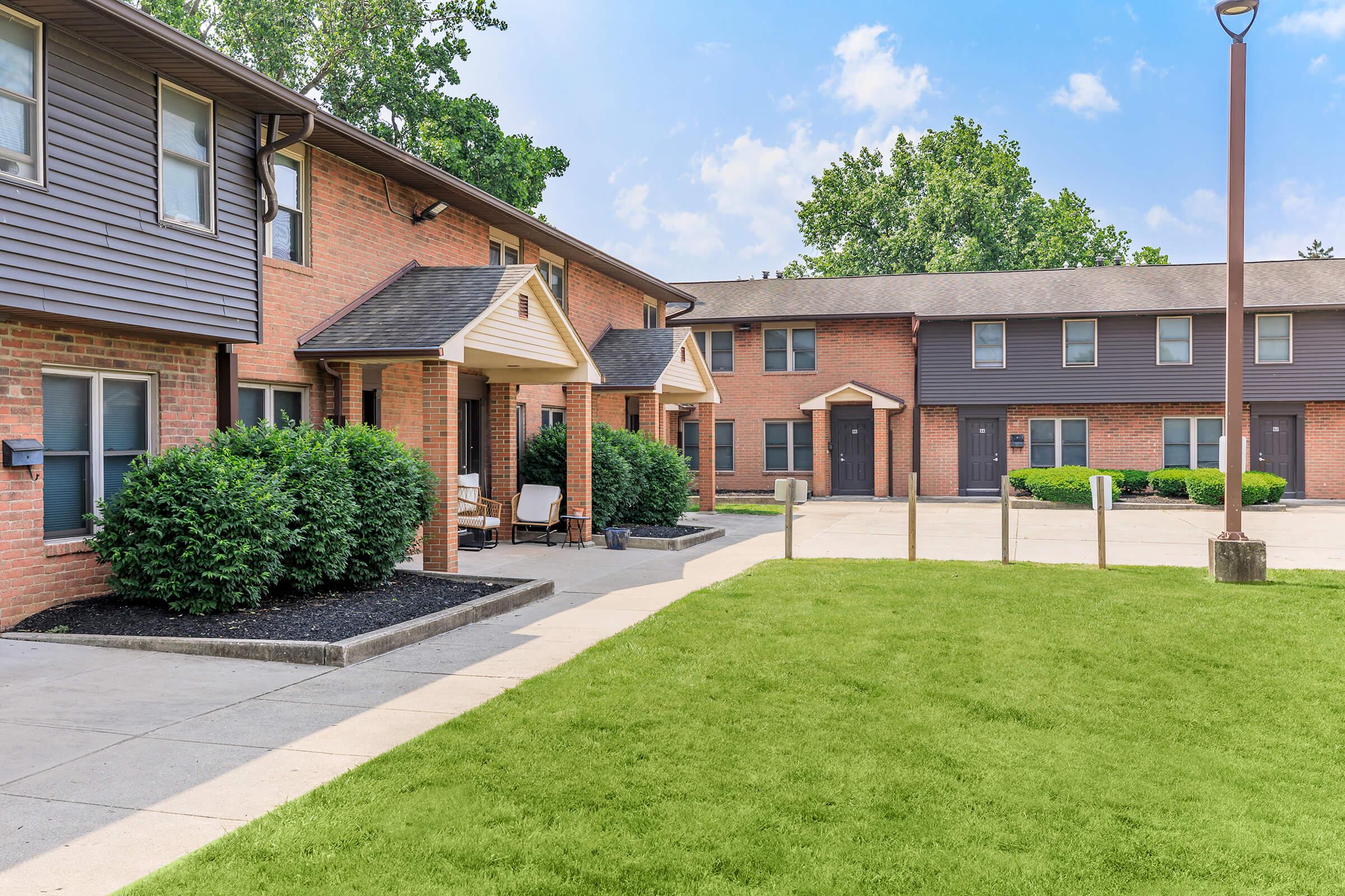 A view of a residential complex featuring two-story brick buildings with gray siding. The foreground includes a well-maintained lawn and pathways leading to the entrances. Bushes surround the doorways, and the background shows additional buildings under a partly cloudy sky.