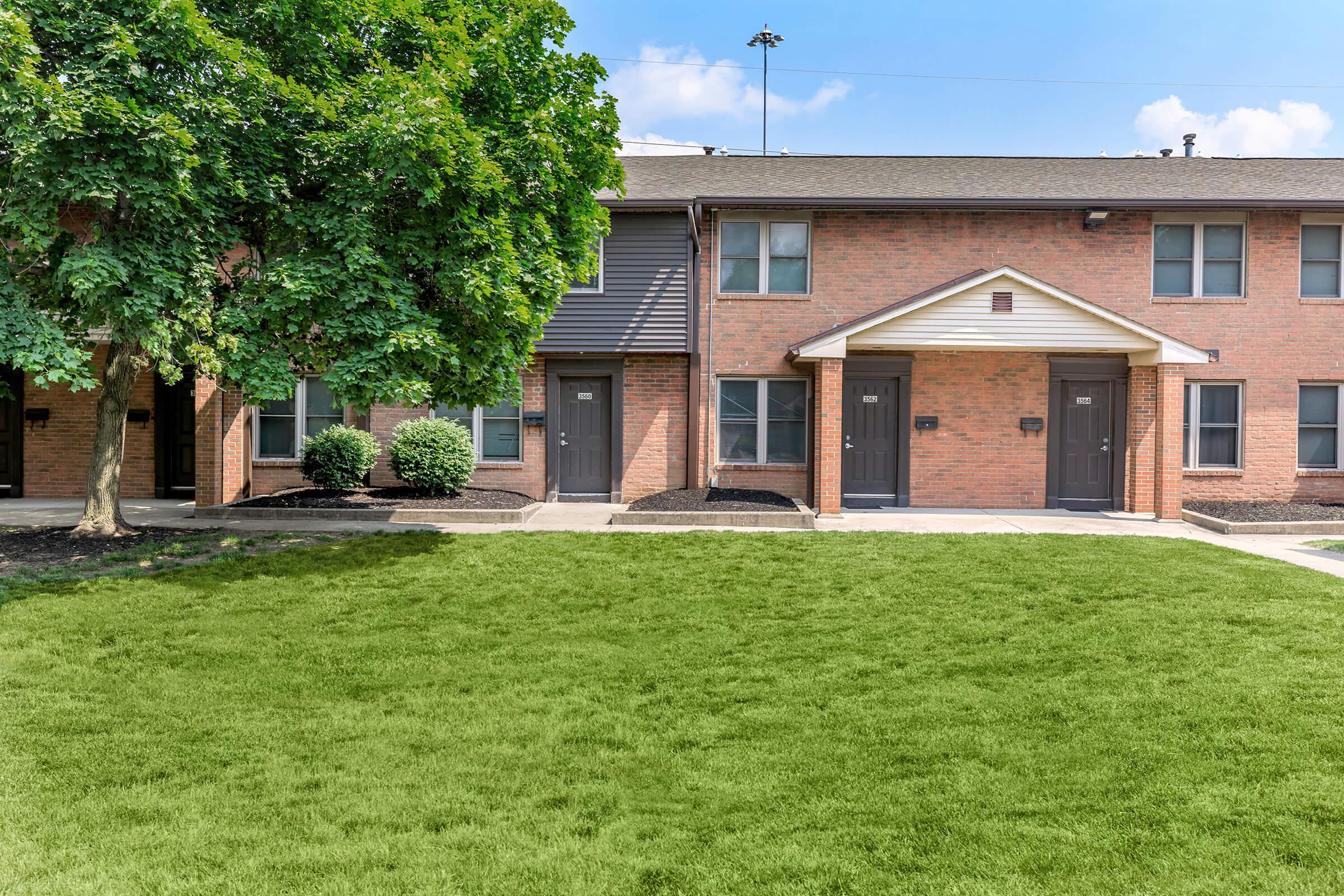 A well-maintained brick apartment building featuring two separate entrances, surrounded by neatly trimmed shrubs and a lush green lawn. A large tree provides shade in the foreground, while clear blue skies are visible above. The overall scene conveys a welcoming residential atmosphere.