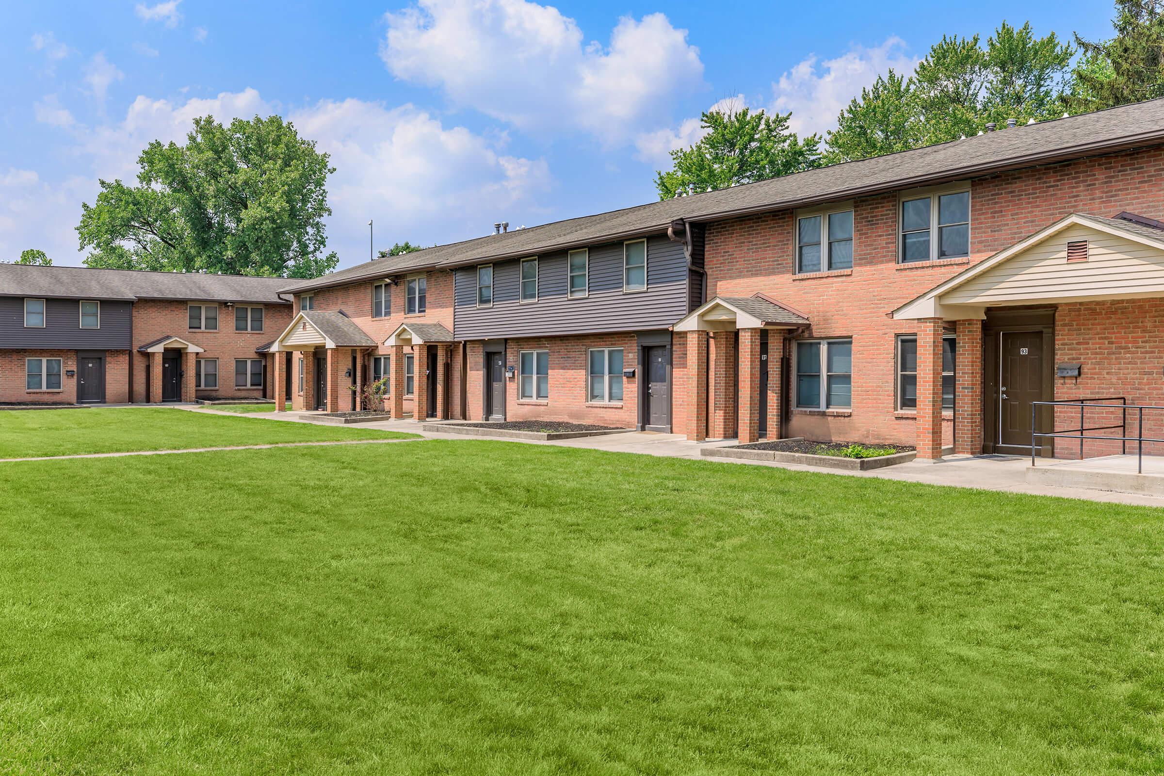A view of a residential building complex featuring multiple two-story brick apartment units. The foreground shows a well-maintained grassy area with walkways leading to the entrances of each unit, surrounded by trees and a clear blue sky with scattered clouds.