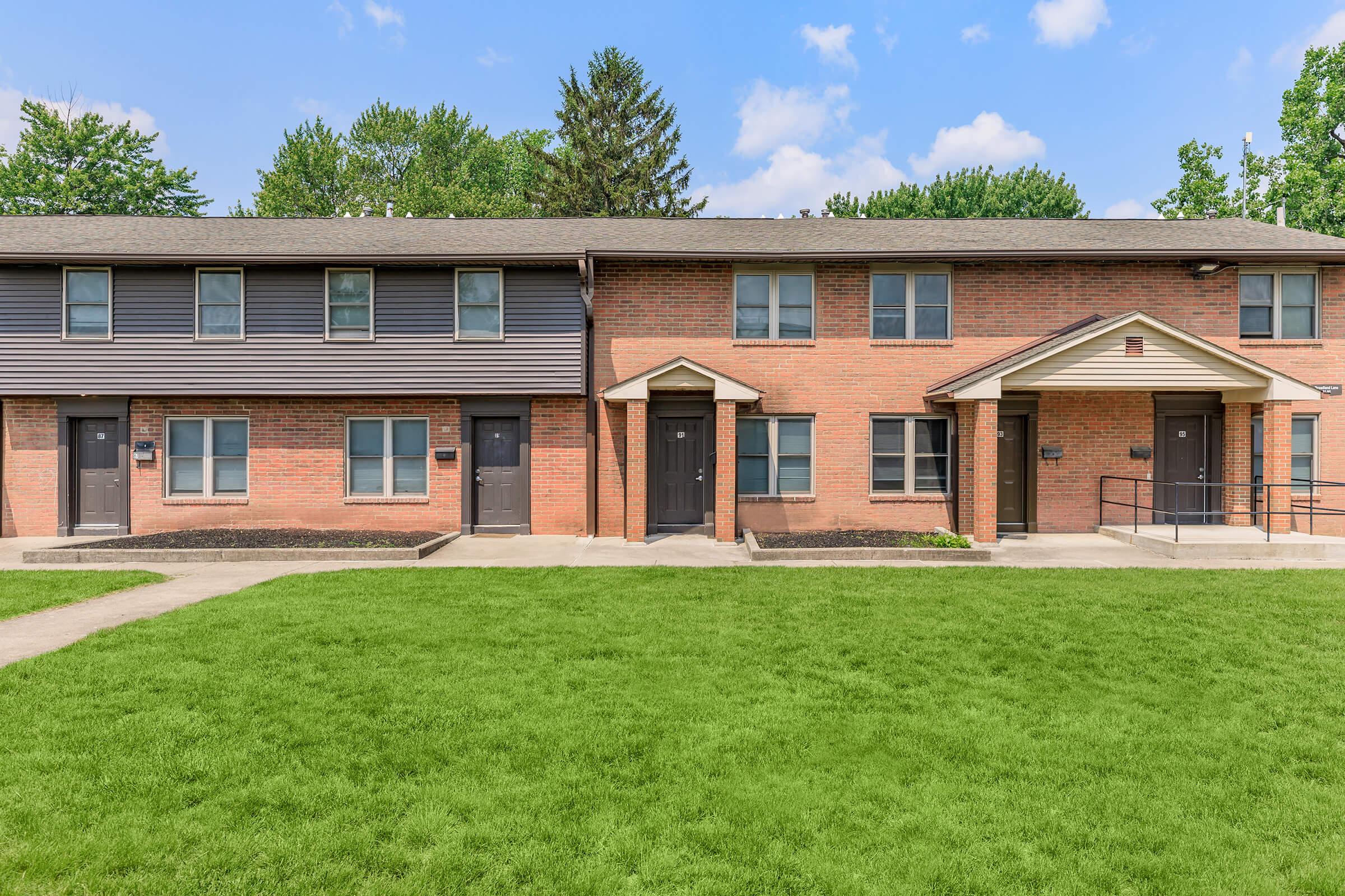 A brick apartment building with two sections, featuring multiple doorways. The left side has a dark, sloped roof, while the right side has a flat roof with an awning above the entrance. A grassy area in front adds a touch of greenery, and trees are visible in the background under a clear blue sky.