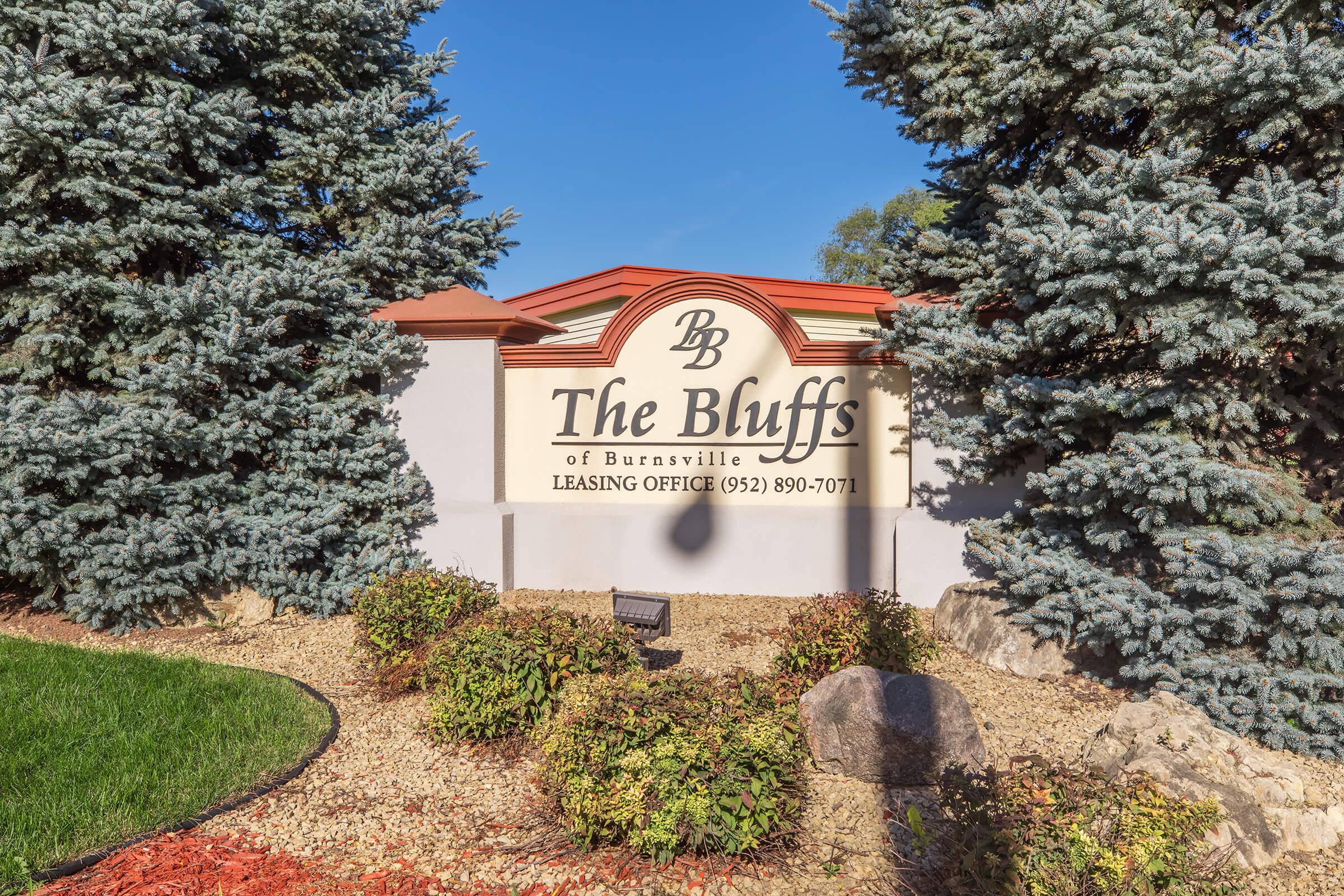 Sign for "The Bluffs of Burnsville" with contact information for the leasing office, surrounded by green grass, colorful shrubs, and conifer trees under a clear blue sky. The area appears well-maintained and inviting.