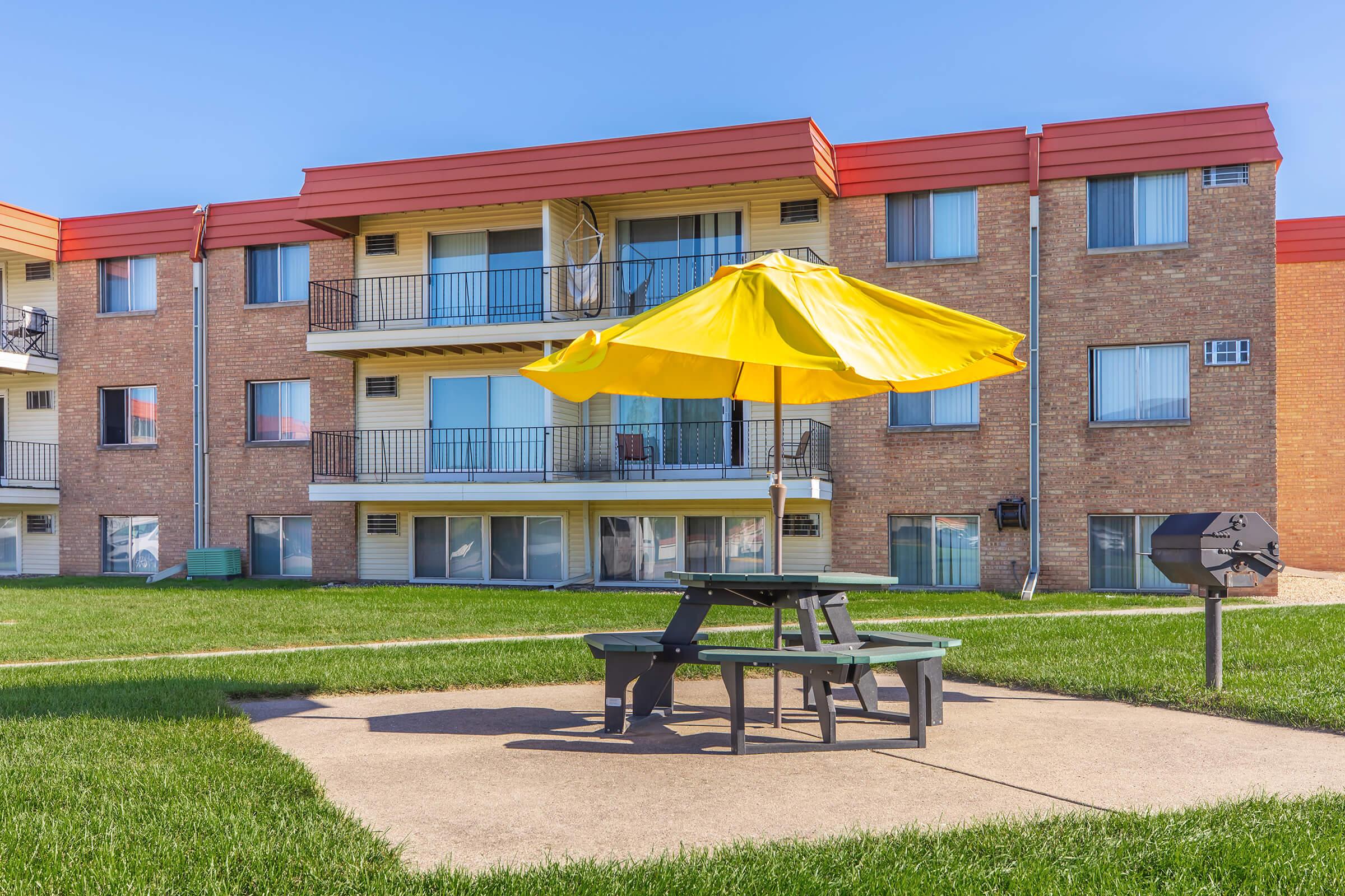 A bright yellow umbrella shades a green picnic table on a concrete pad, surrounded by lush grass. In the background, a two-story brick apartment building features multiple balconies with sliding glass doors and colorful roofs, under a clear blue sky.