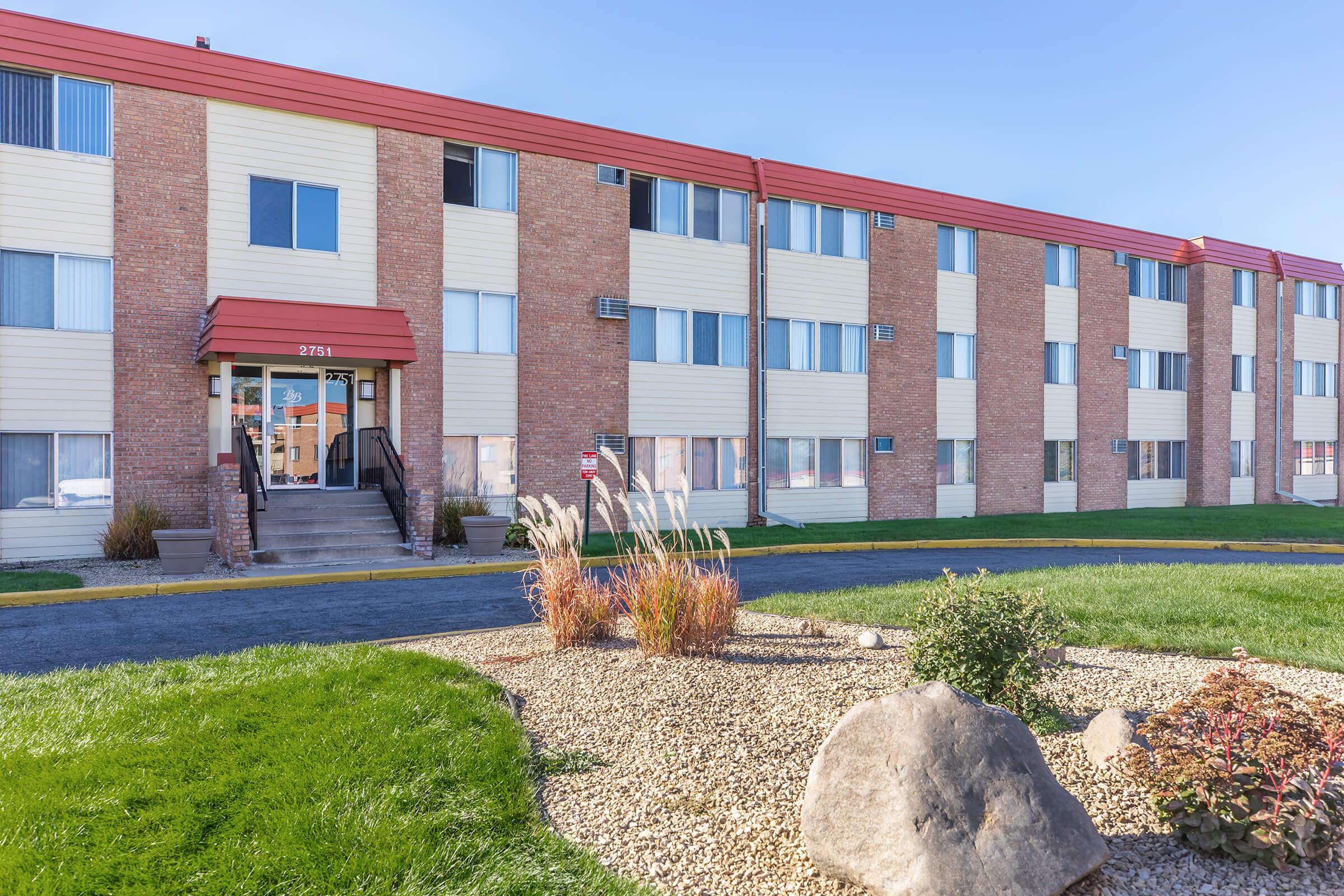 A three-story apartment building with a brick exterior and cream-colored panels. The entrance features a covered awning and stairs leading up to the door. Surrounding the building is a well-maintained lawn with decorative shrubs and rocks, under a clear blue sky.