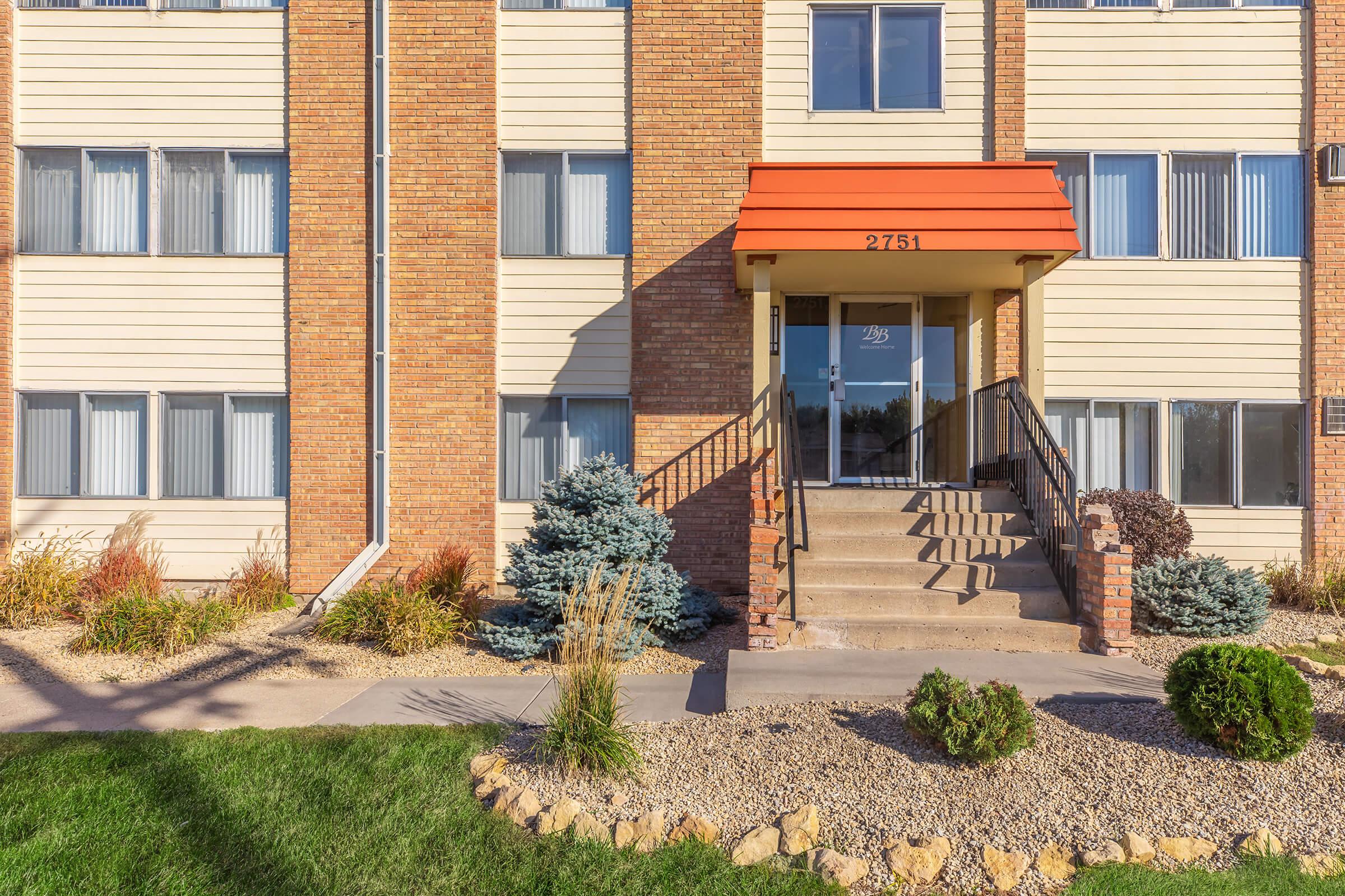 Entrance of an apartment building featuring a brick façade, large windows, and a prominent porch with steps. The door is framed by a red awning. Lush landscaping with shrubs and ornamental grasses surrounds the entryway, along with a gravel path leading to the entrance.