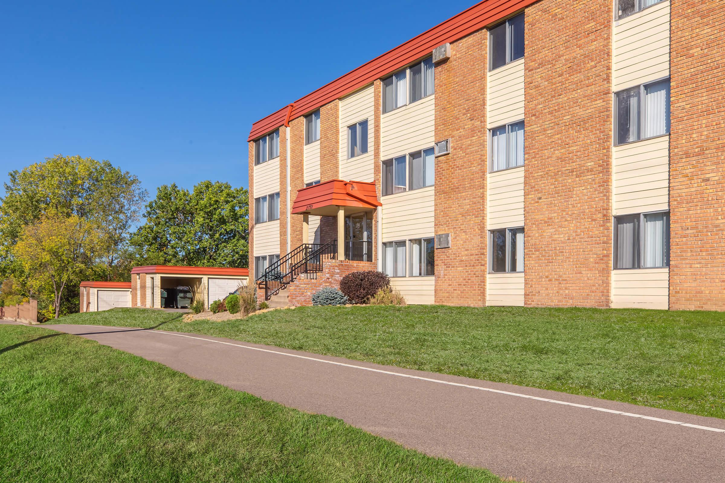A multi-story residential building with a brick and siding exterior. The entrance features a covered porch with stairs leading up. Lush green grass surrounds the building, and a paved driveway runs alongside it. Trees and blue sky are visible in the background, suggesting a clear, sunny day.