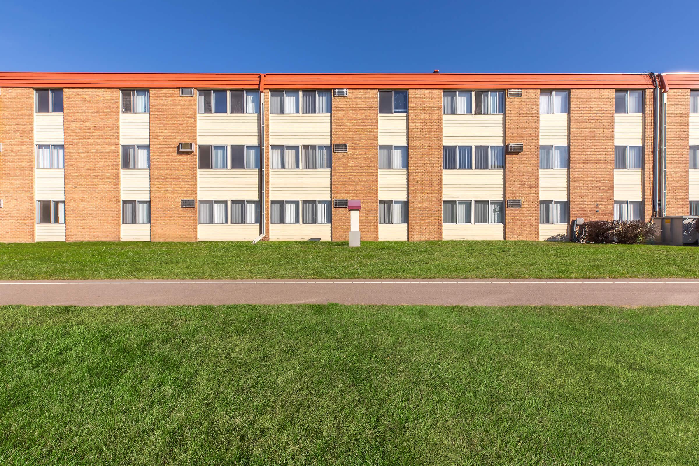 A low-rise apartment building with a red and beige facade, featuring multiple windows and air conditioning units. The grassy area in the foreground is well-maintained, and a paved path runs alongside the building. Bright blue sky overhead adds to the vibrant scene.