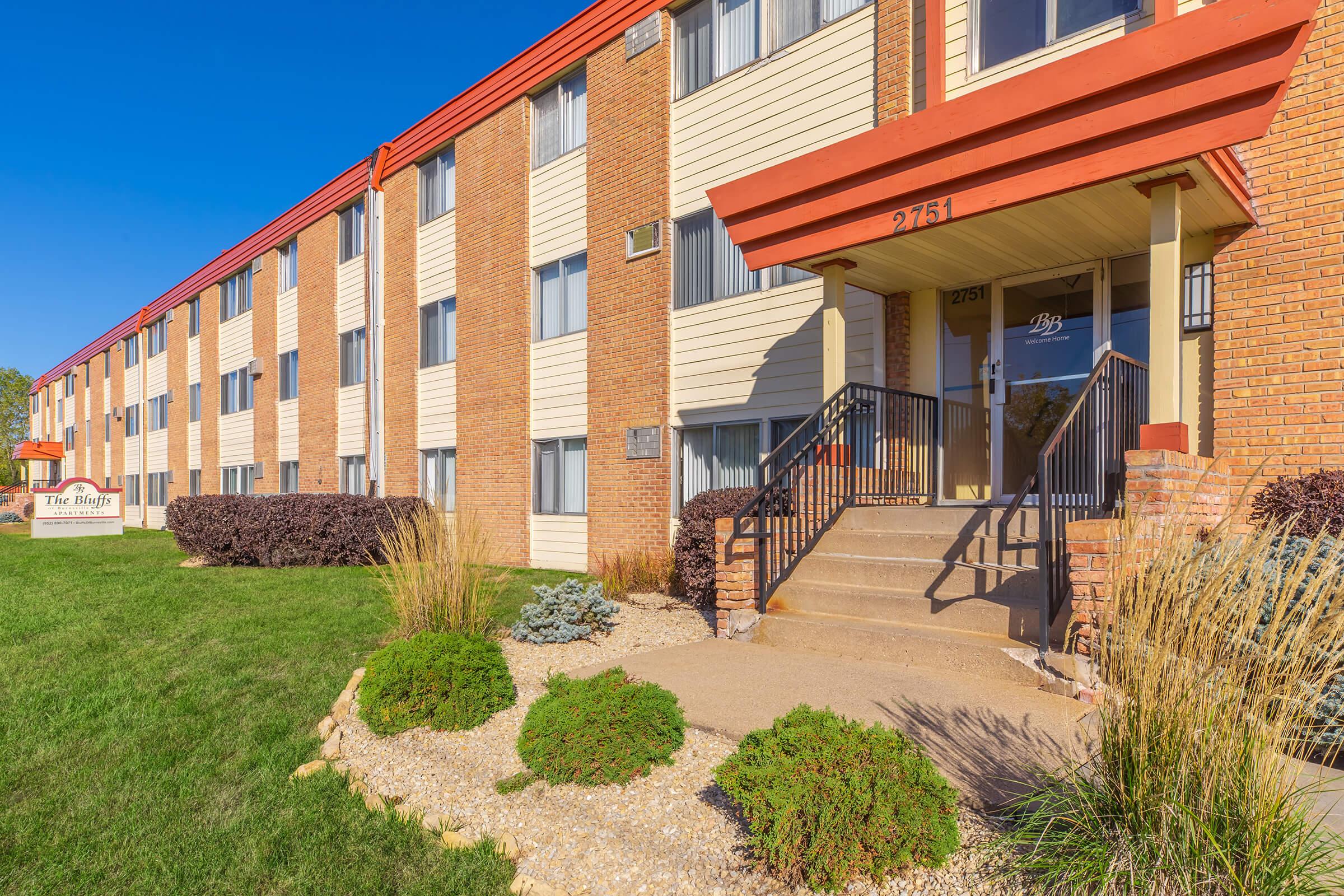 A three-story apartment building with a brick and beige exterior. The entrance features a set of steps leading up to the door, framed by red awnings. Lush green grass and low shrubs surround the entrance, with a sign in front displaying the building's name. Clear blue sky overhead.