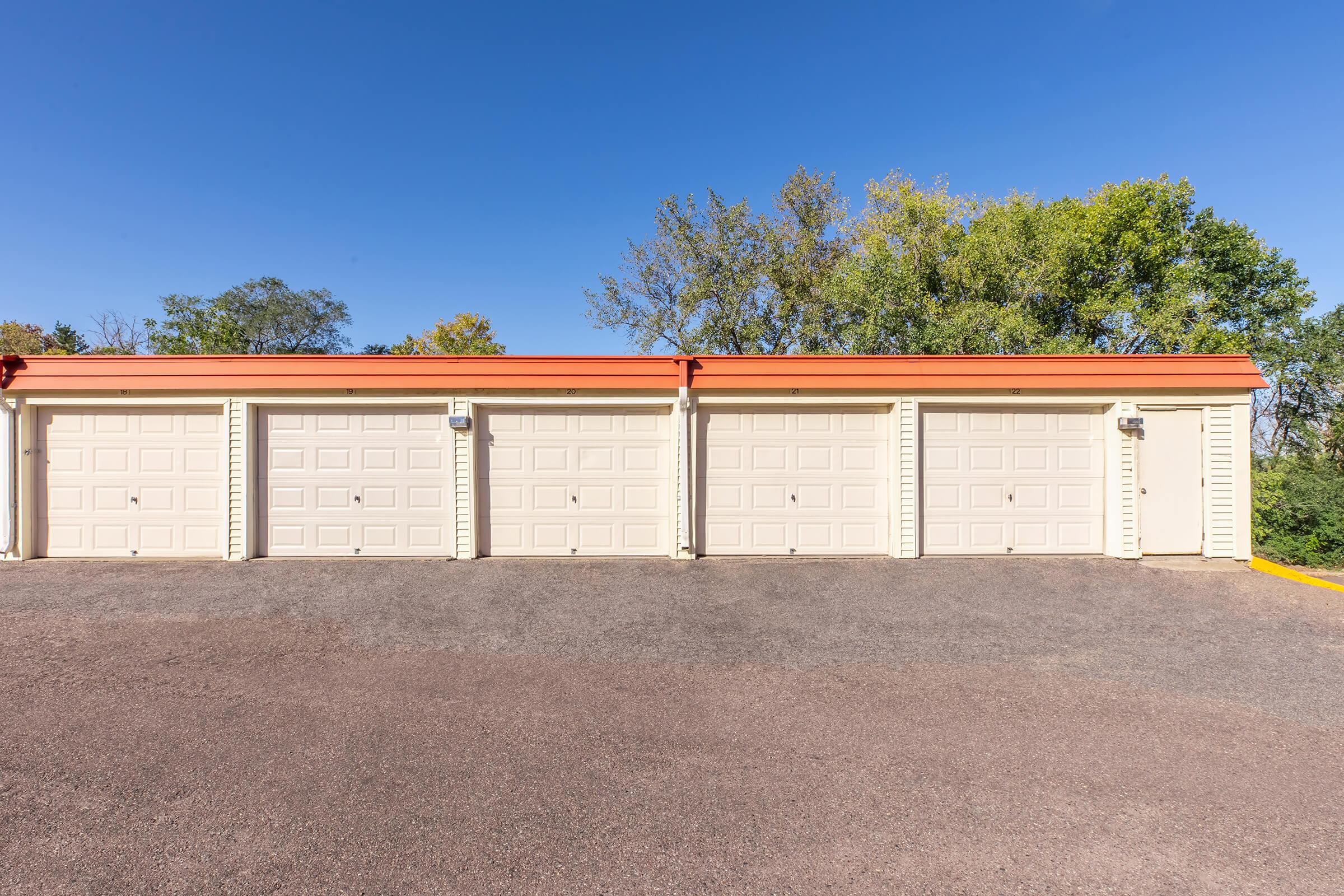 A row of beige storage units with orange roofs, set against a clear blue sky. The paved driveway in front is clear and well-maintained, with green trees visible in the background. The storage units are closed and feature simple, panelled doors.