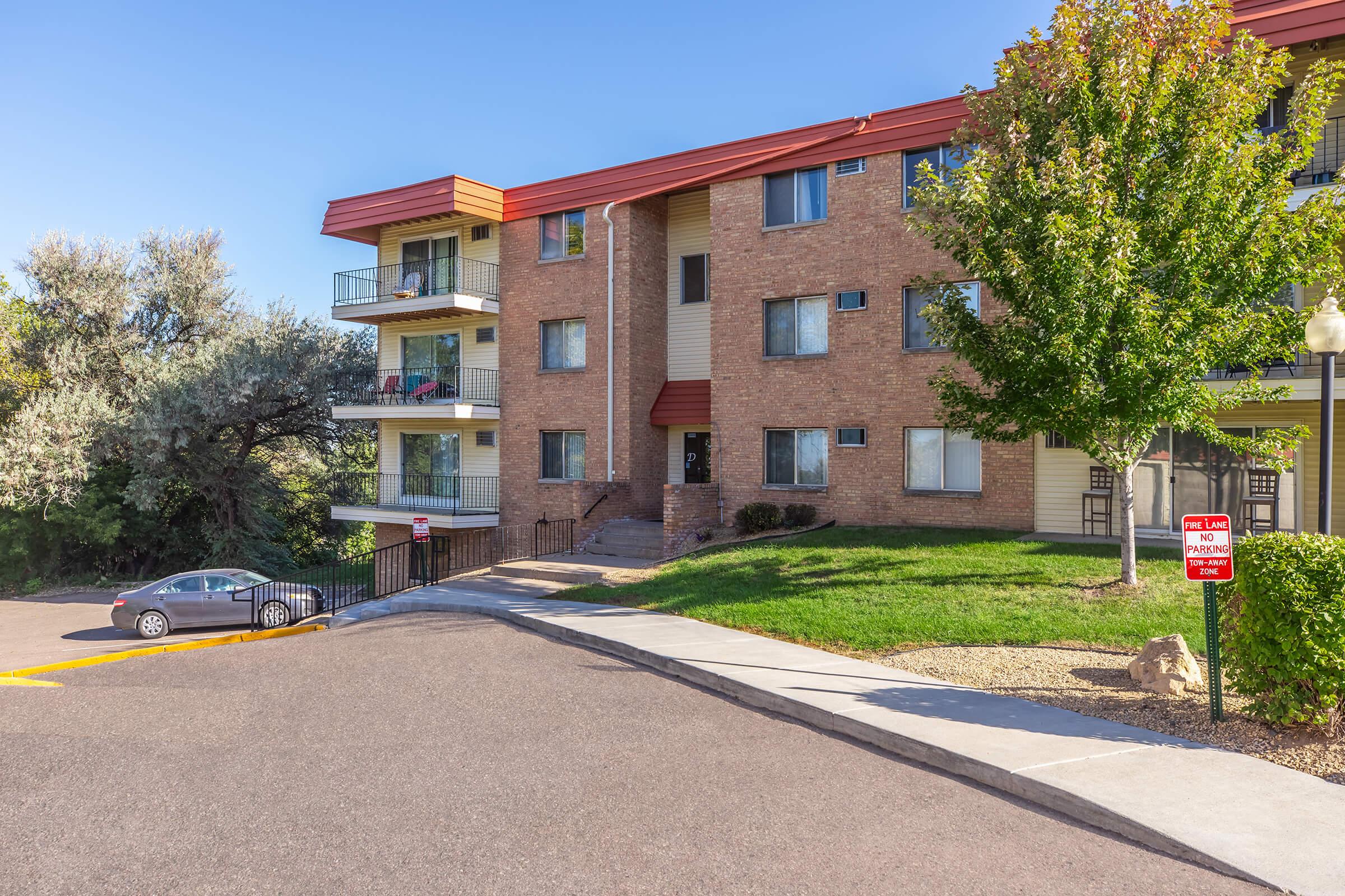 A multi-story brick apartment building with red accents, featuring multiple balconies. The entrance has steps leading up from a parking area. Surrounding greenery includes trees and shrubs, under a clear blue sky. A sign indicating parking regulations is visible near the entrance.