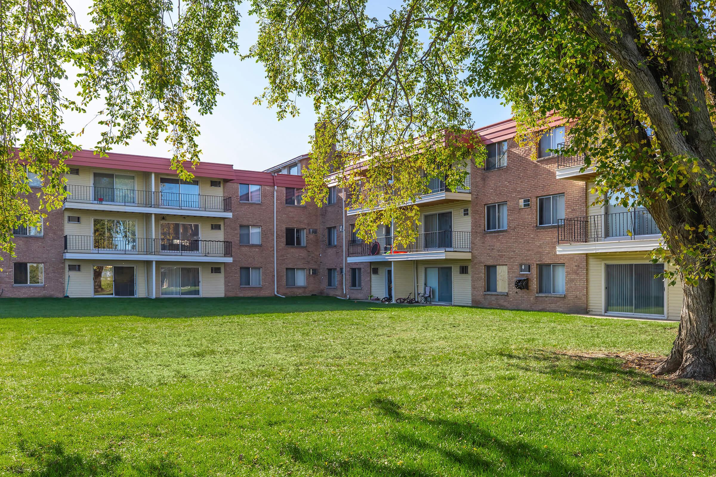 A view of a residential apartment complex featuring several brick buildings with balconies. The lush green lawn in the foreground is framed by trees. The sky is clear and sunny, creating a warm atmosphere for the outdoor space.