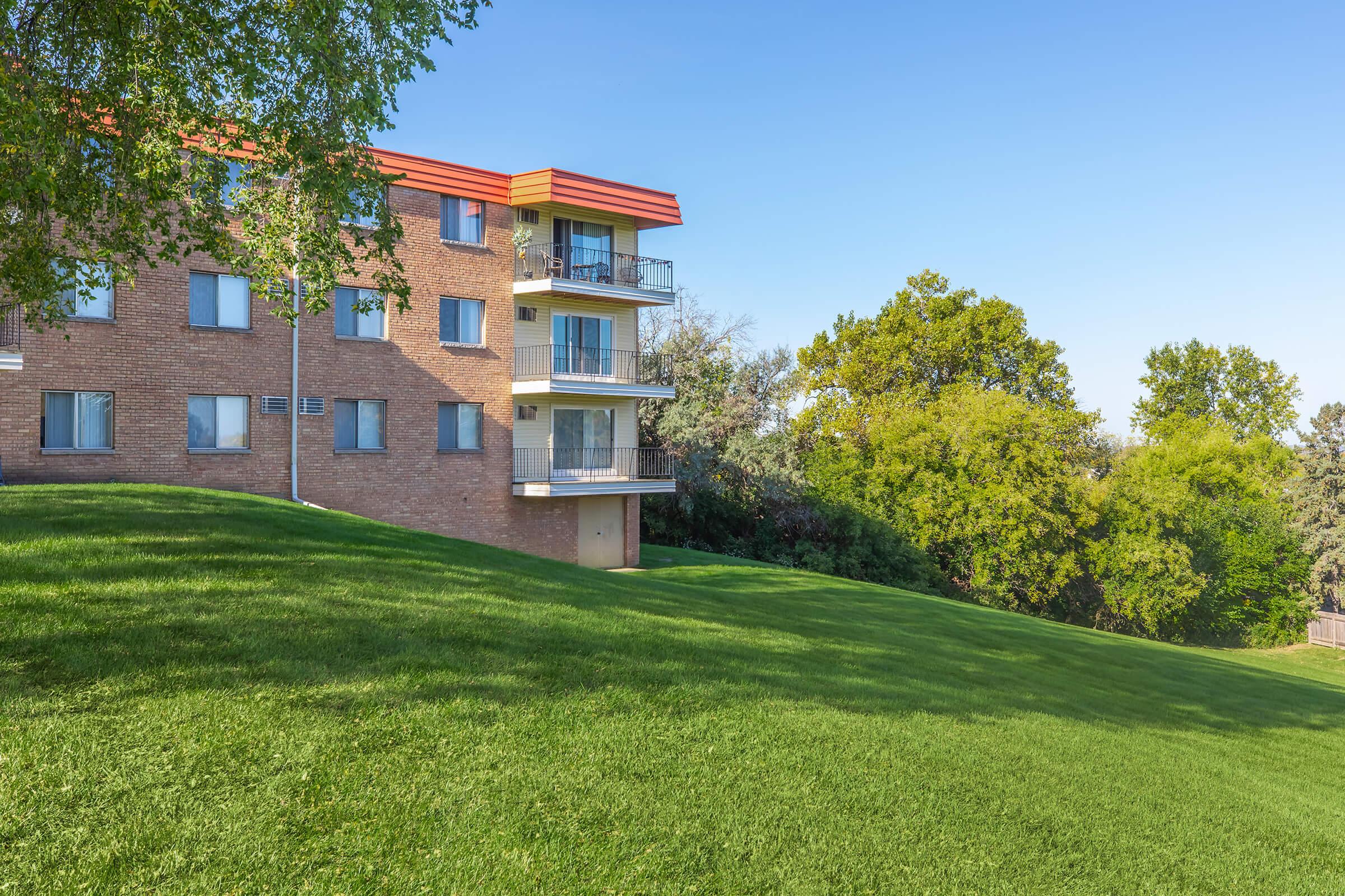 A multi-story brick apartment building with a modern red roof, situated on a gently sloping green lawn. Surrounding the building are trees and shrubs, and the sky is clear with no clouds. The scene conveys a peaceful, suburban atmosphere.