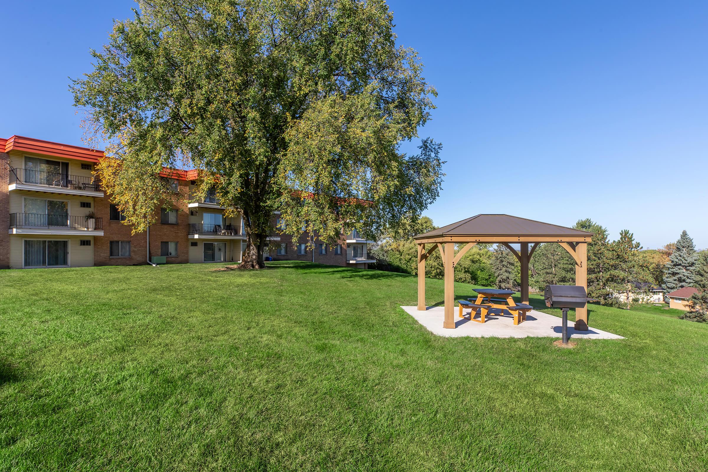 A spacious green lawn featuring a wooden gazebo with a picnic table and grill, surrounded by trees. In the background, there are two multi-story apartment buildings with balconies, under a clear blue sky.