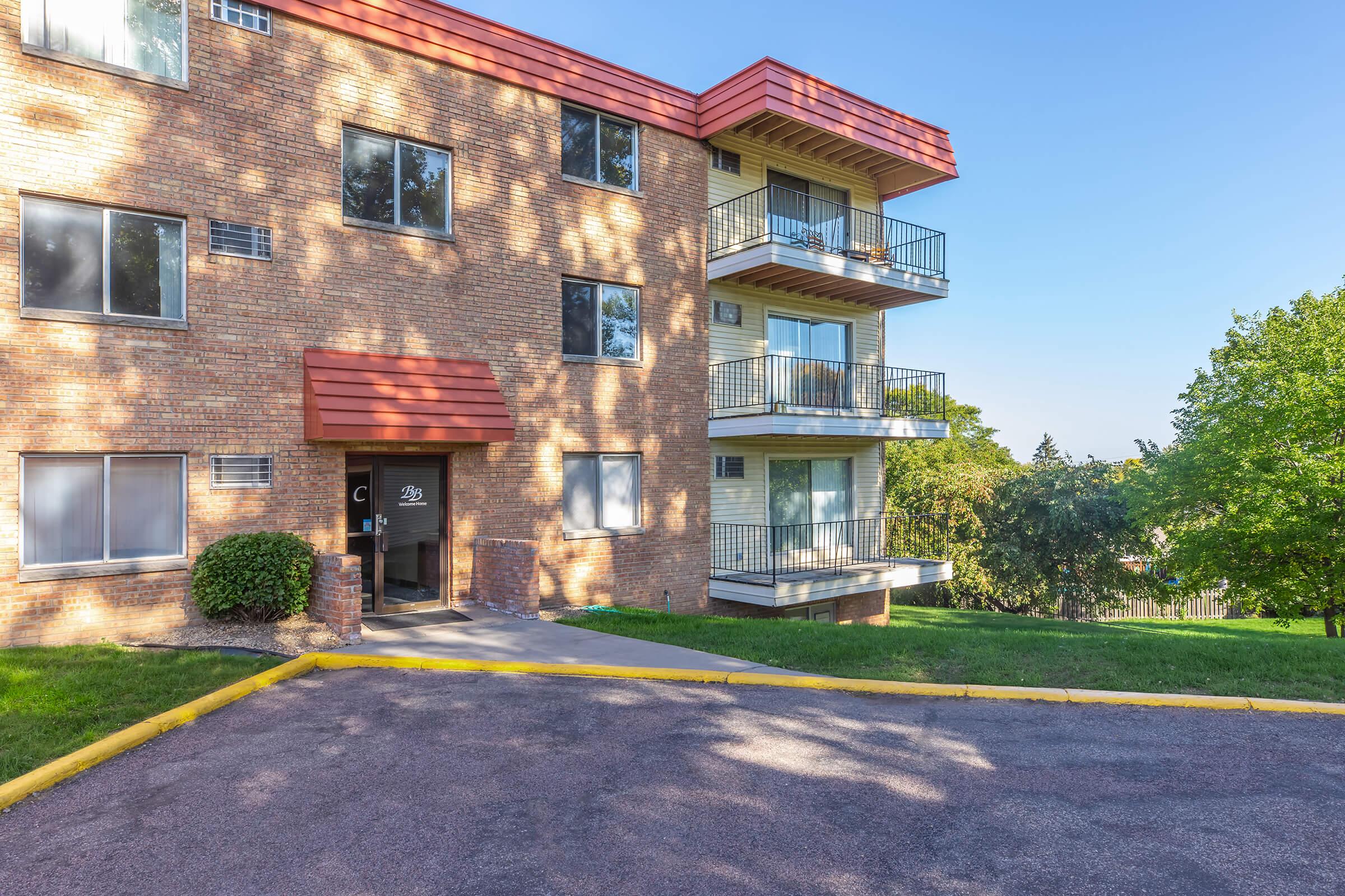 A multi-story brick apartment building with a red roof. The facade features large windows and balconies on the upper levels. A landscaped area with trees and grass is visible in the foreground, along with a paved walkway leading to the entrance. The sky is clear and blue, indicating a sunny day.