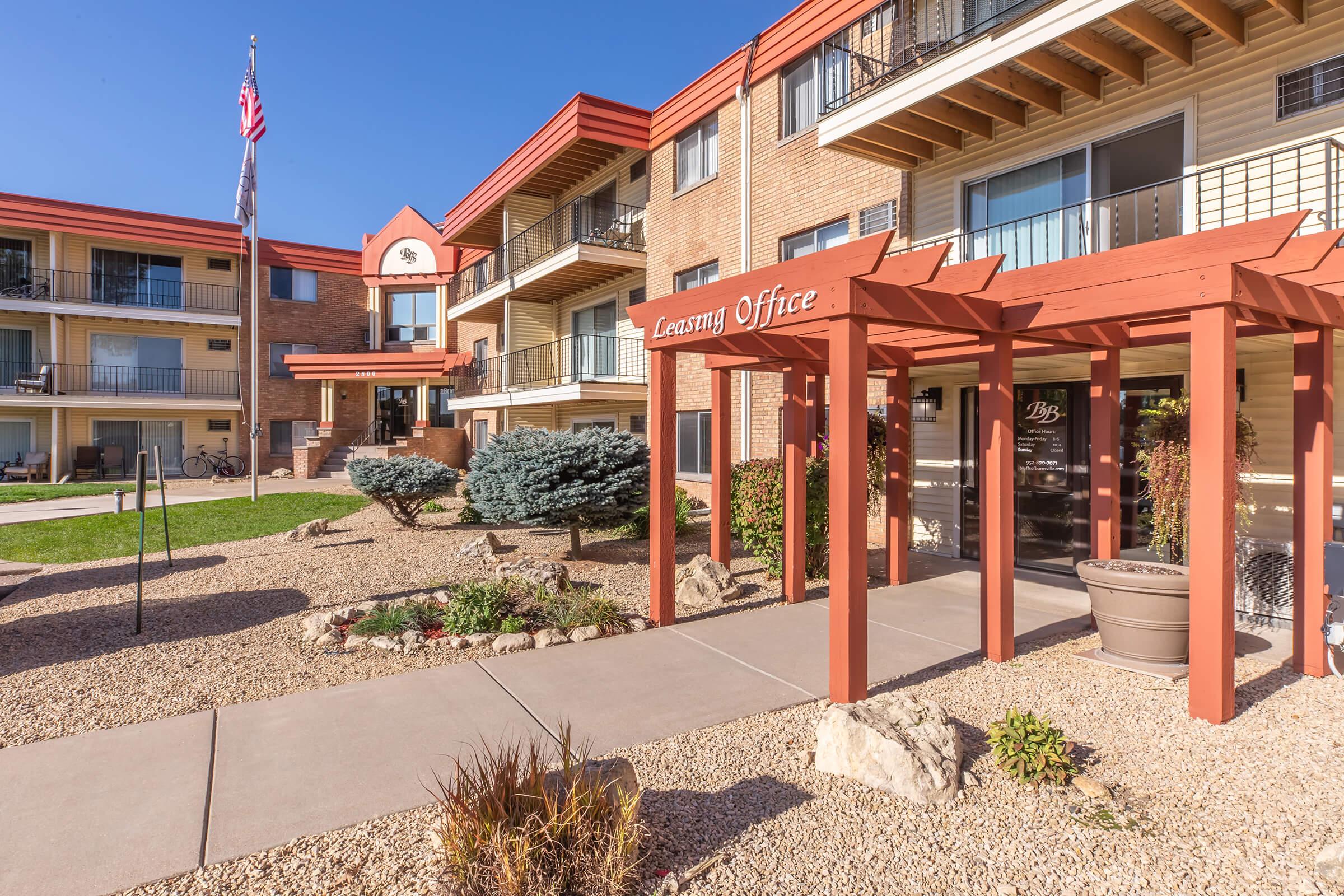 A well-maintained apartment complex featuring a leasing office with a wooden overhang, surrounded by landscaped areas with bushes and rocks. The building has multiple balconies and an American flag displayed, under a clear blue sky, creating a welcoming atmosphere.