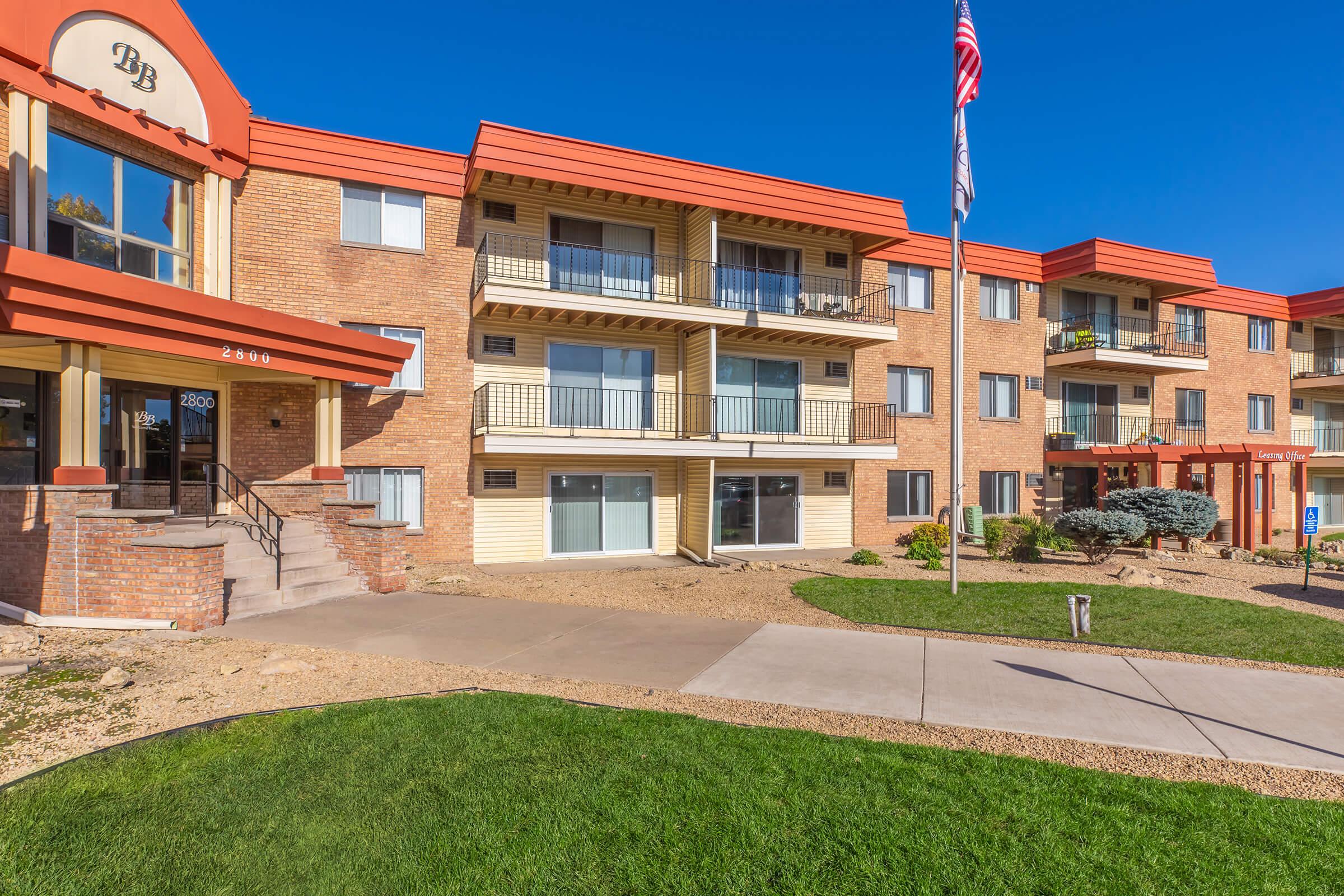 A modern residential building featuring multiple balconies and a brick facade. The structure has a welcoming entrance with stairs, manicured lawns, and decorative landscaping. An American flag is prominently displayed in front of the building, with clear blue skies in the background.
