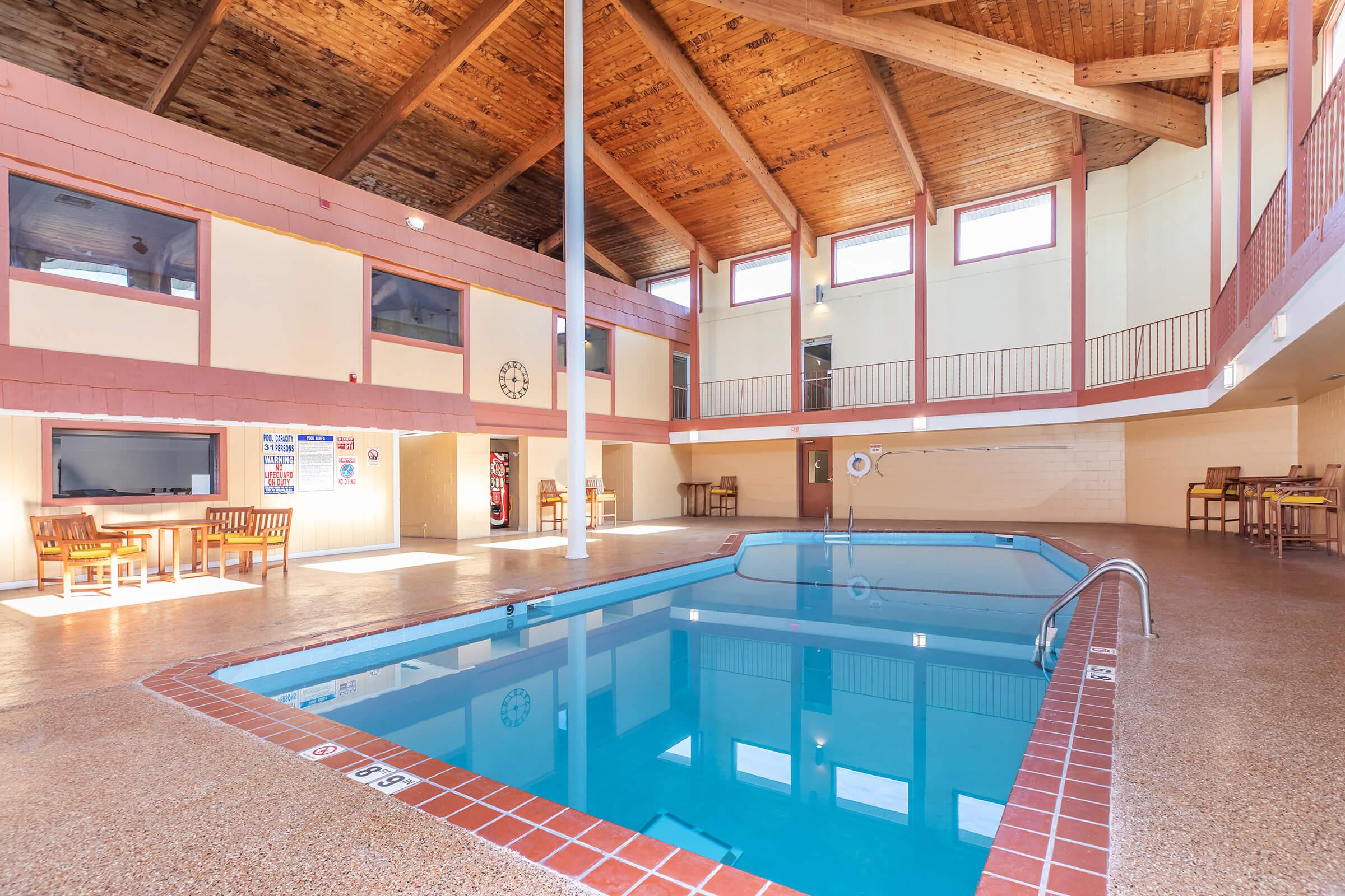 Indoor pool area featuring a rectangular pool with a shallow end, surrounded by a tiled deck. Natural wood beams support the high ceiling, with large windows allowing sunlight to brighten the space. There are tables and chairs nearby, creating a relaxed atmosphere. A vending machine is also visible.
