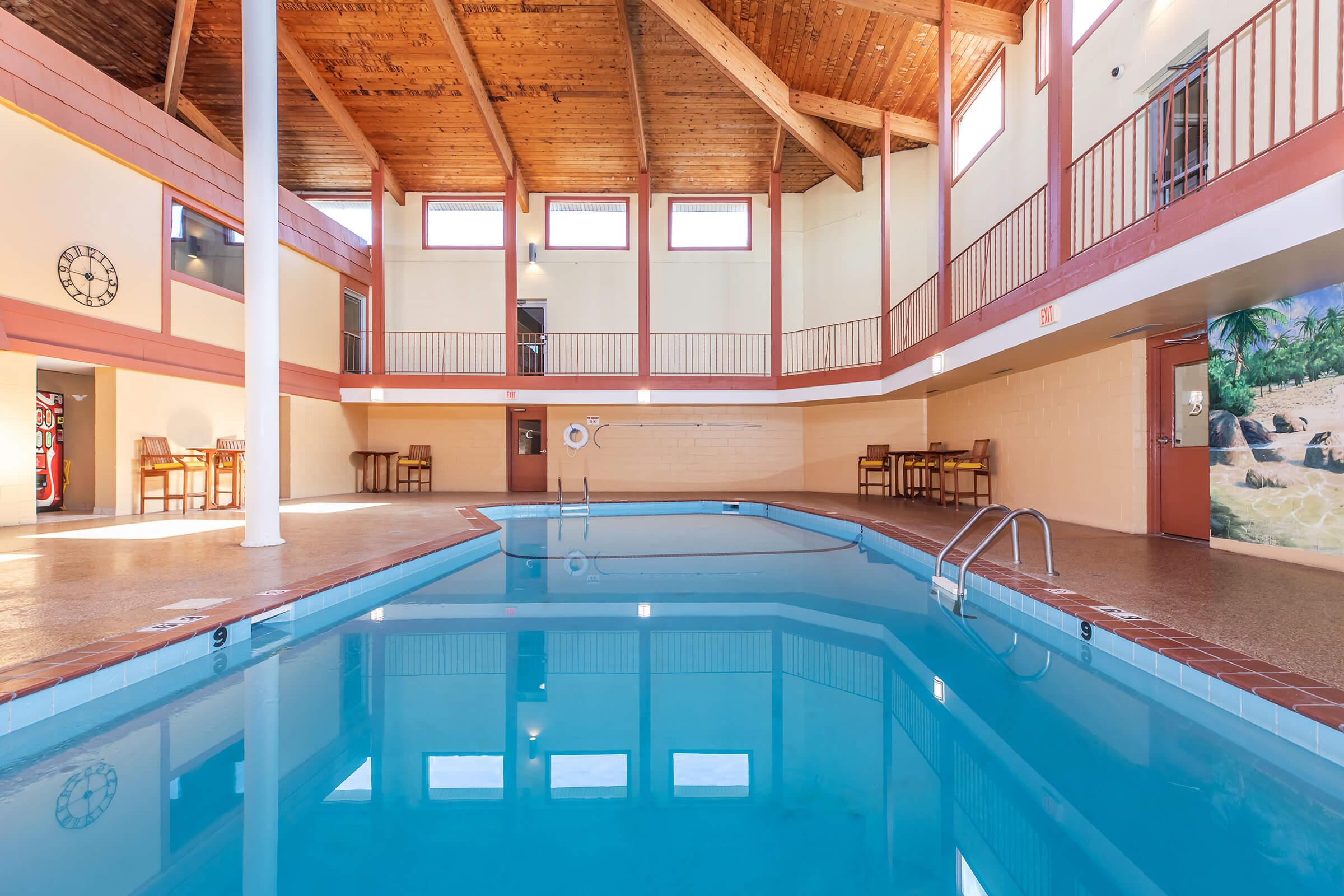 Indoor swimming pool area featuring clear blue water, wooden beam ceilings, and large windows for natural light. Surrounding space includes a few chairs, a lifebuoy on the wall, and a mural depicting a tropical scene. The overall ambiance is bright and inviting.