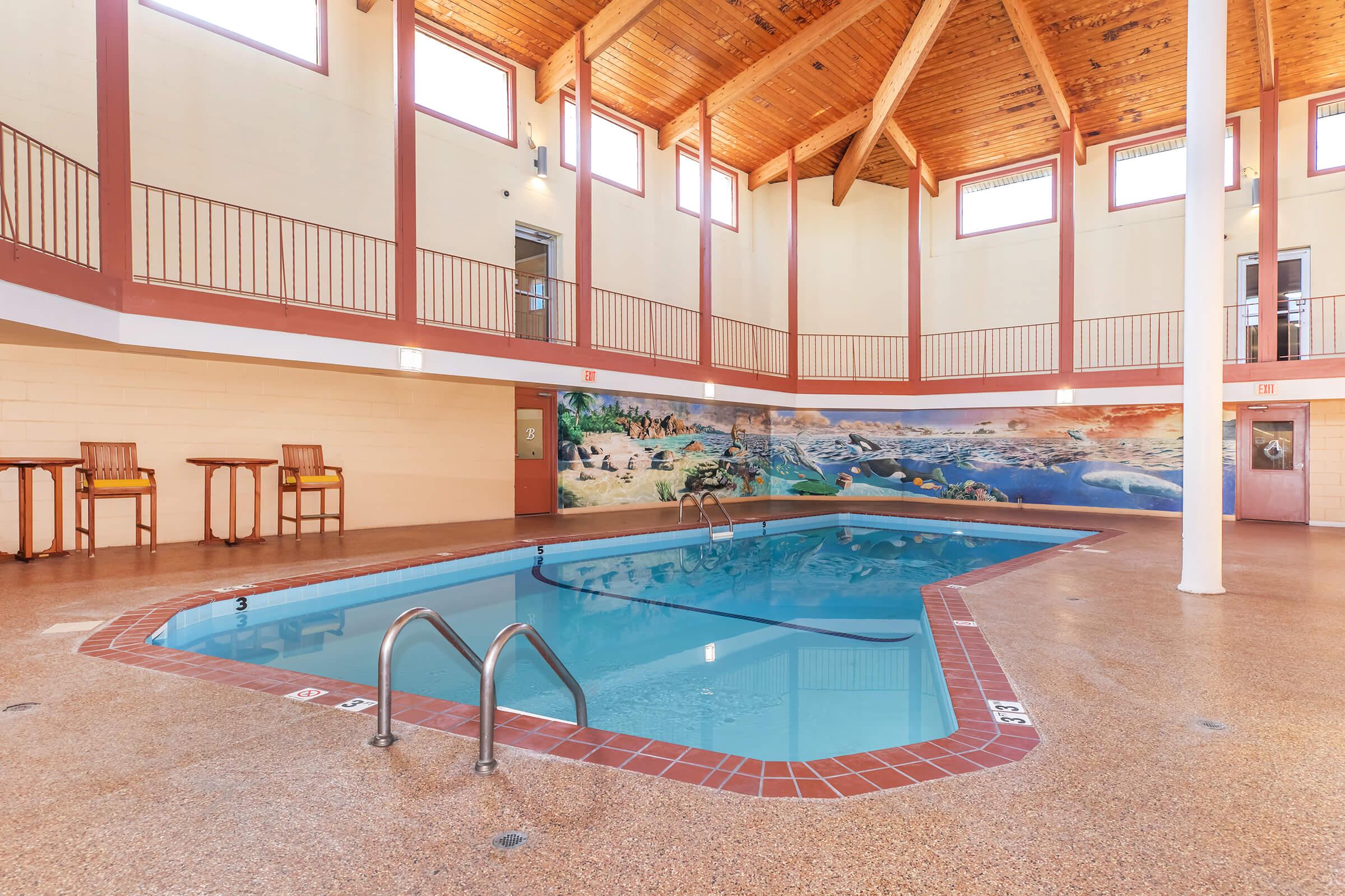 Indoor swimming pool with clear blue water, surrounded by light-colored walls and large windows letting in natural light. The ceiling features wooden beams, and there are a few chairs positioned nearby. A mural of underwater scenery decorates one wall, enhancing the relaxing atmosphere.