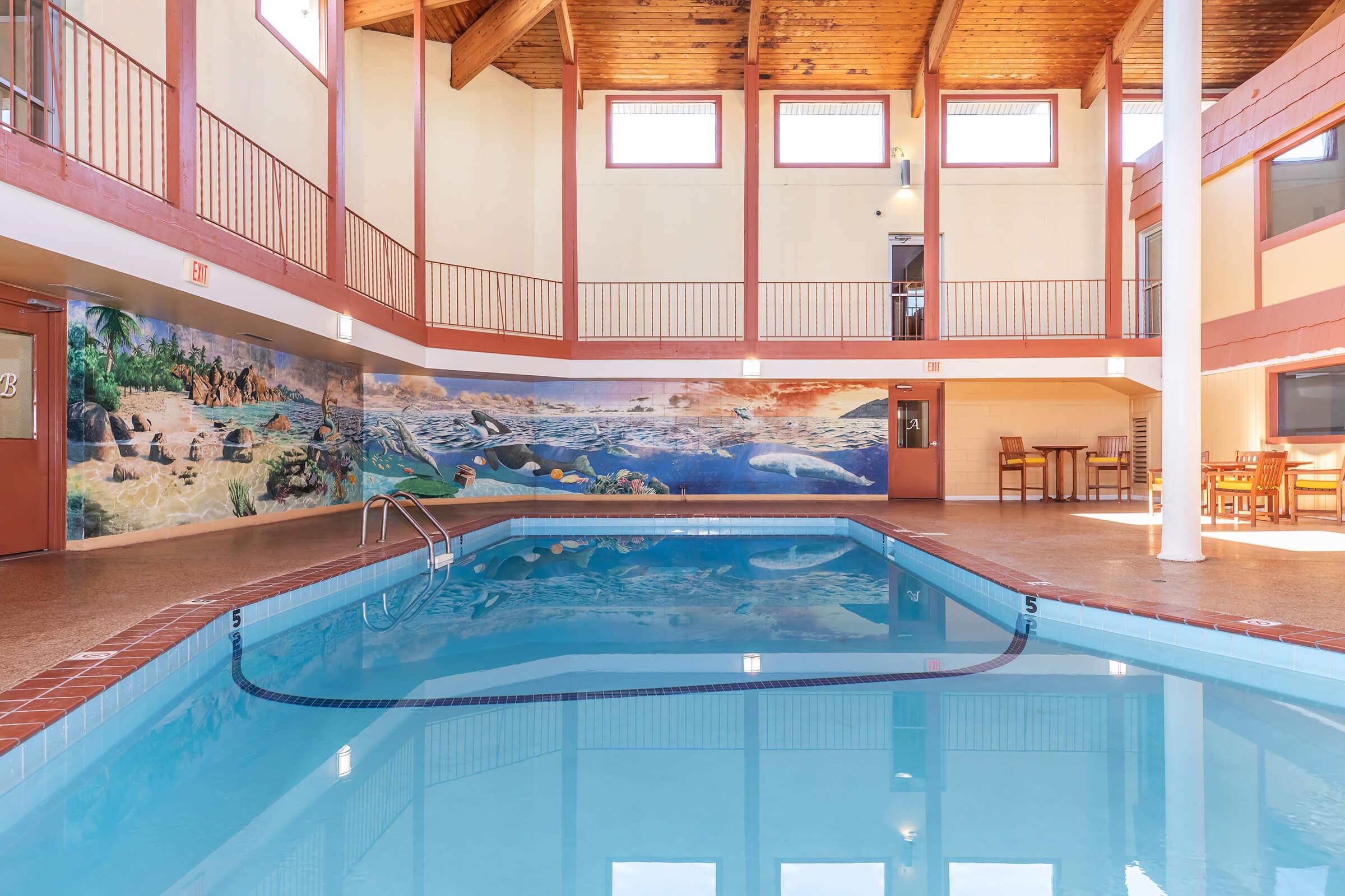 Indoor swimming pool with clear blue water, surrounded by wooden beams and vibrant murals depicting marine life on one wall. Tables and chairs are situated nearby, creating a welcoming atmosphere for relaxation and leisure. Natural light fills the space through large windows.