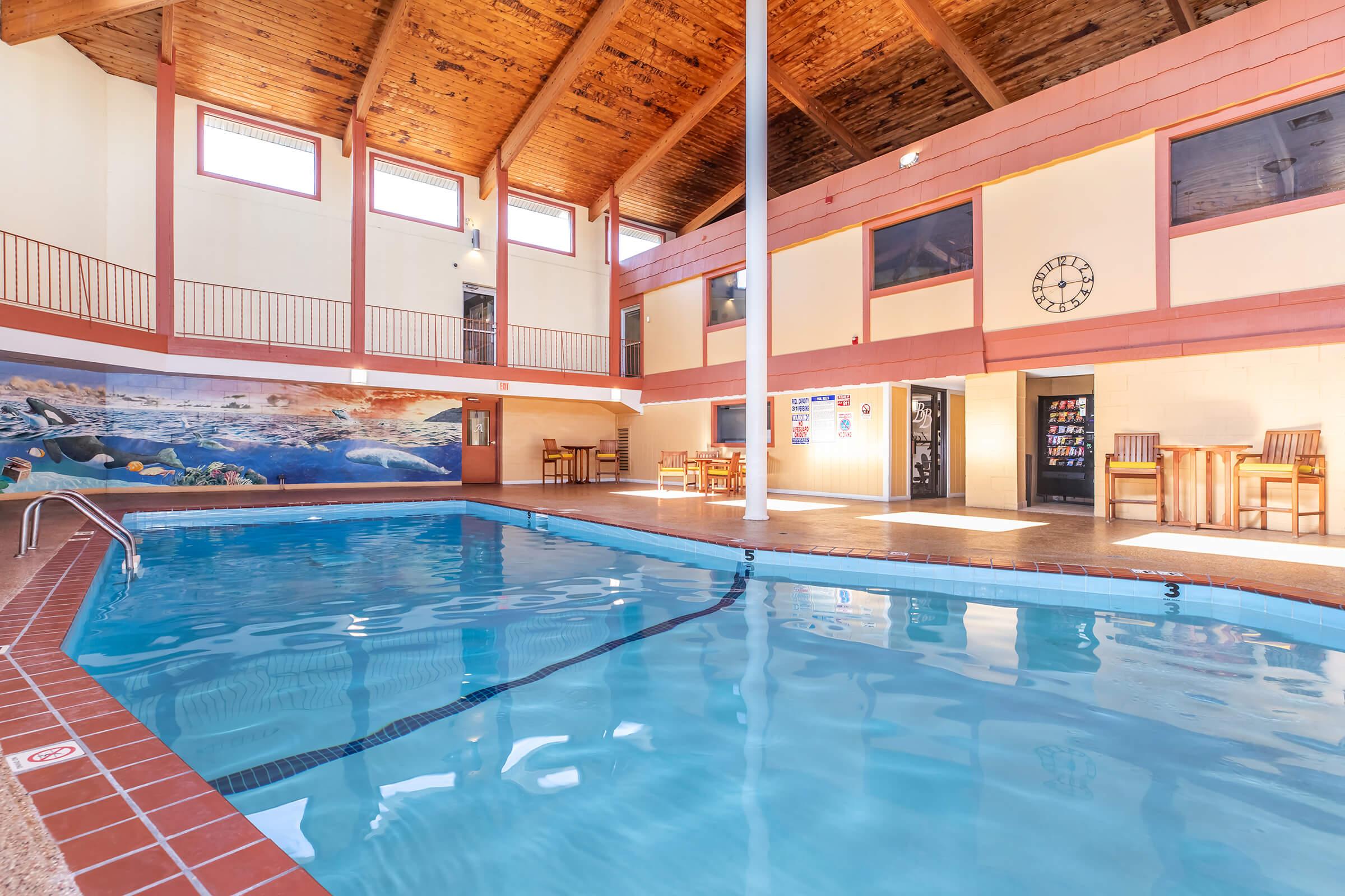 Indoor swimming pool with a spacious area featuring a wooden ceiling. The pool is surrounded by tiled flooring and has a mural of an underwater scene on the wall. Tables and chairs are set up nearby, and vending machines can be seen in the background. Large windows allow natural light to fill the space.