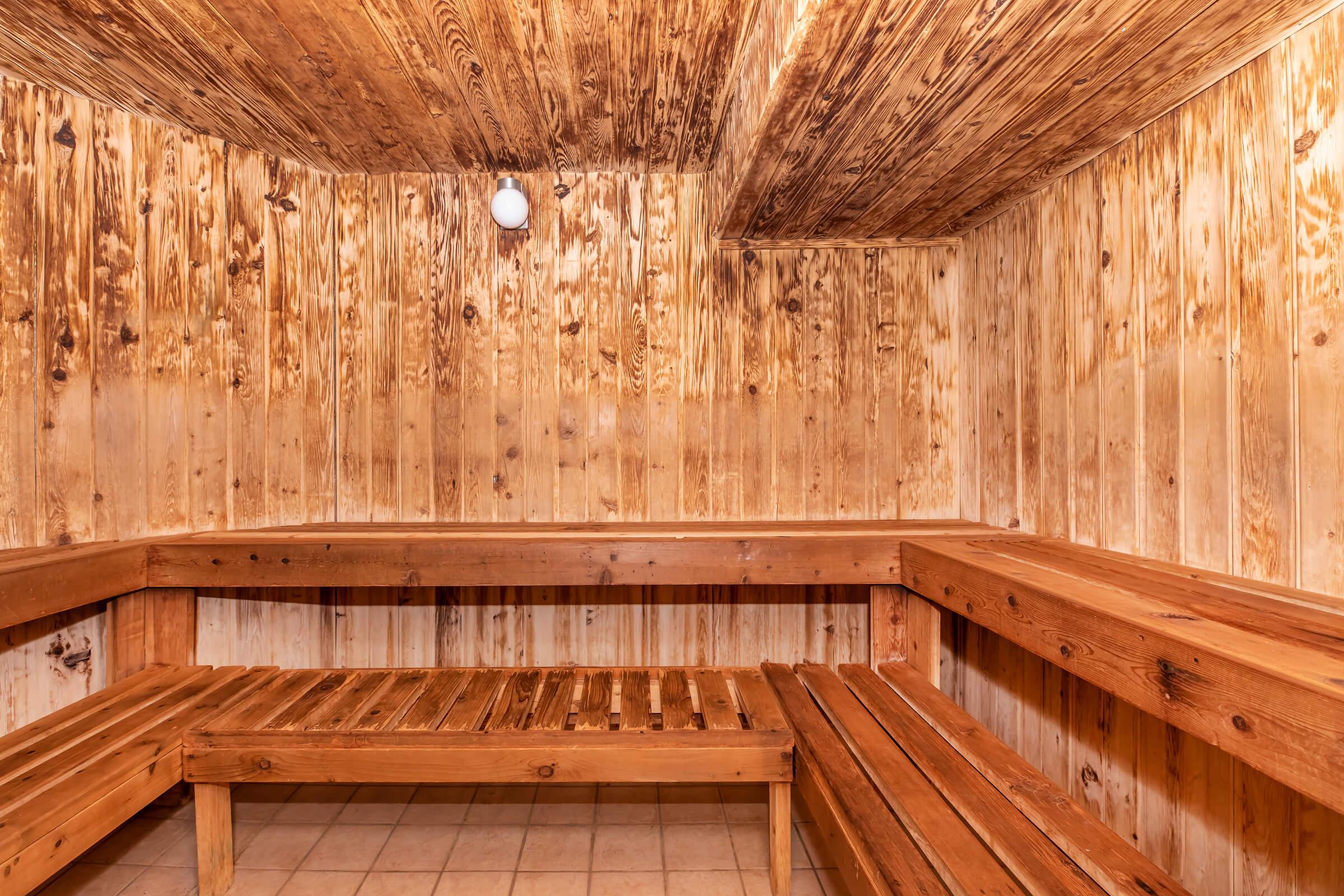 Interior of a wooden sauna featuring bench seating made of slatted wood. The walls and ceiling are adorned with light-colored wooden panels, creating a warm and inviting atmosphere. A single light bulb is mounted on the ceiling, providing illumination in the space. The floor is tiled.